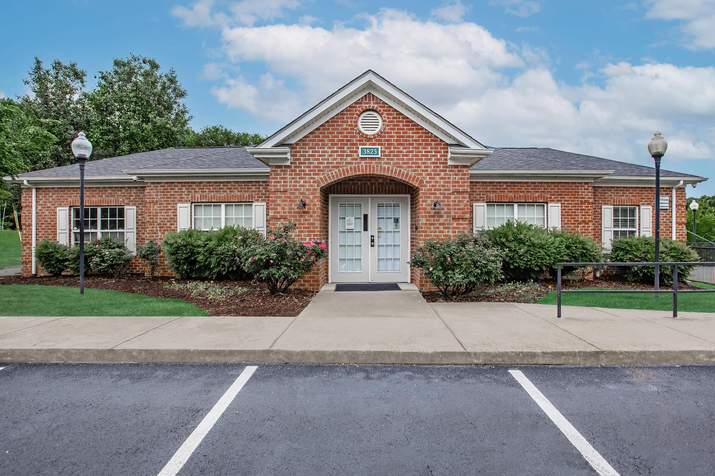 A red brick building with white trim and a front porch, surrounded by shrubs and landscaping. The sky is partly cloudy, and there is a parking lot in front. The building appears to be well-maintained, with decorative lampposts on either side of the walkway.
