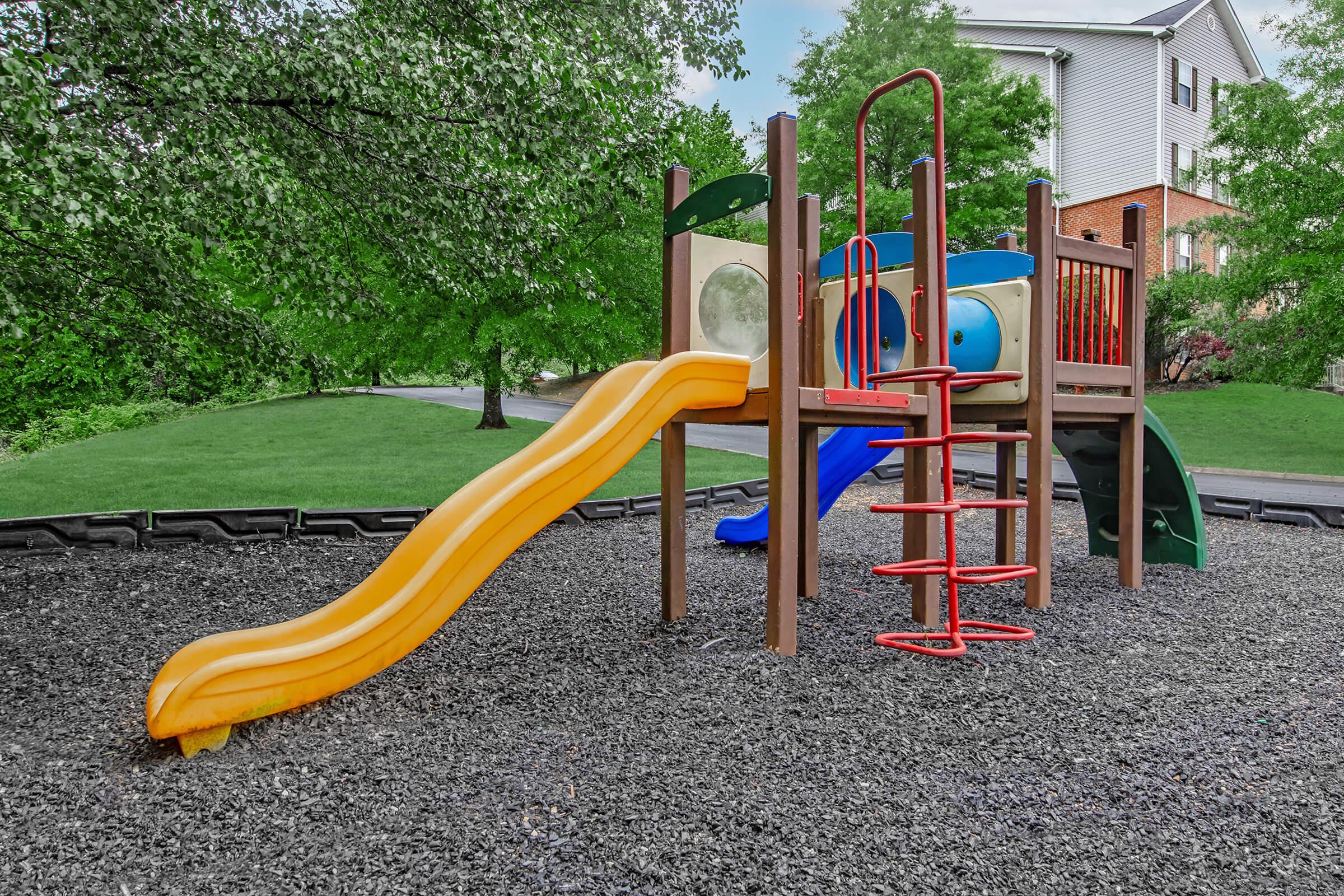 A colorful playground structure with a yellow slide, climbing features, and a blue element. The playground is set in a green, grassy area surrounded by trees and a pathway, providing a cheerful space for children to play.