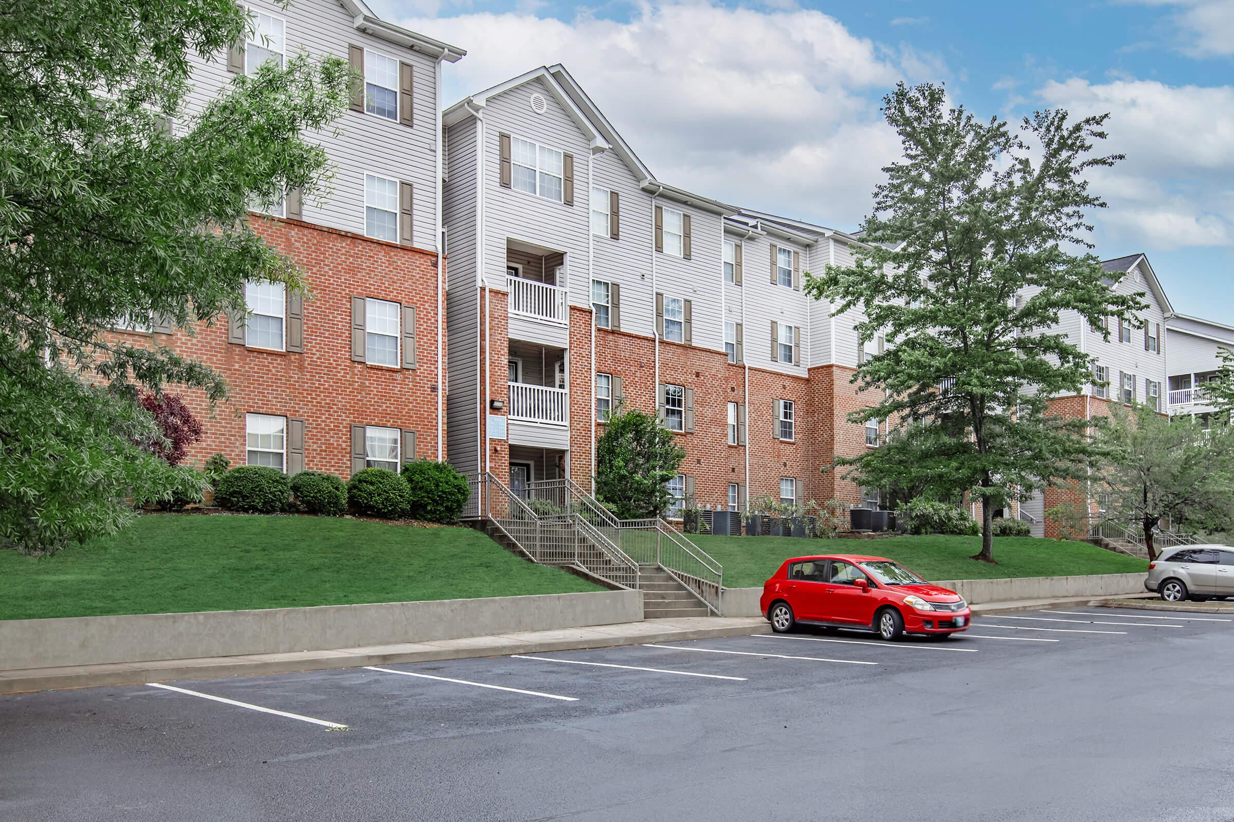 A residential apartment building with a brick facade and multiple levels. The building features balconies and is surrounded by green lawns and trees. In the foreground, there is a parking lot with a red car parked on the left and additional parked vehicles in the background. The sky is partly cloudy.
