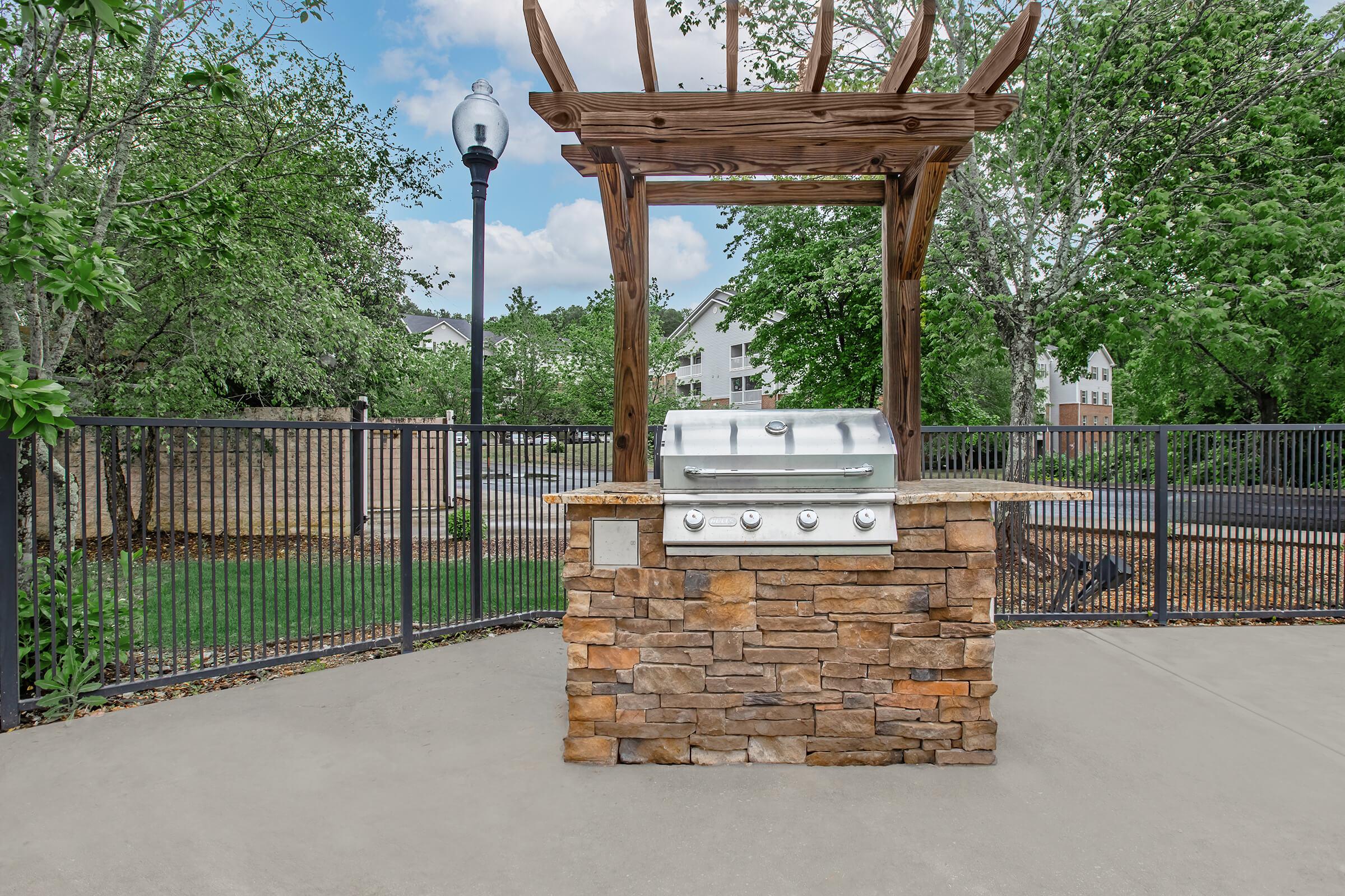 Outdoor barbecue grill built into a stone structure under a wooden pergola. Surrounded by a fenced area with green trees and grass in the background, creating a pleasant outdoor cooking space. The scene is bright and inviting, ideal for entertaining.
