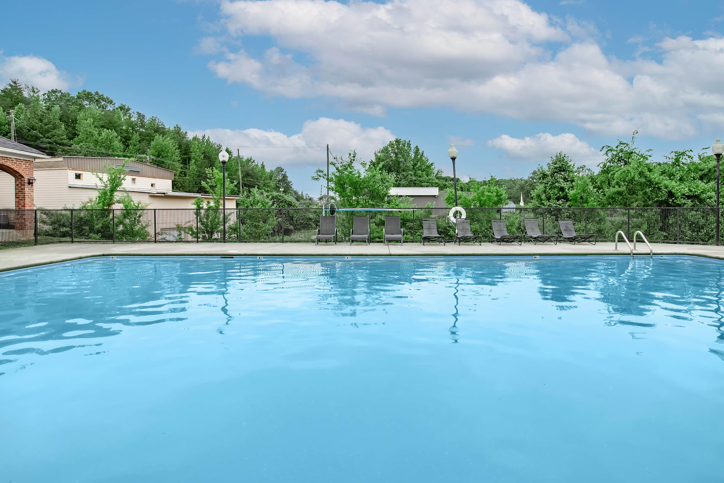 A clear blue swimming pool surrounded by greenery and lounge chairs. The sky is partly cloudy, creating a serene atmosphere. Trees and buildings are visible in the background, adding to the tranquil setting.