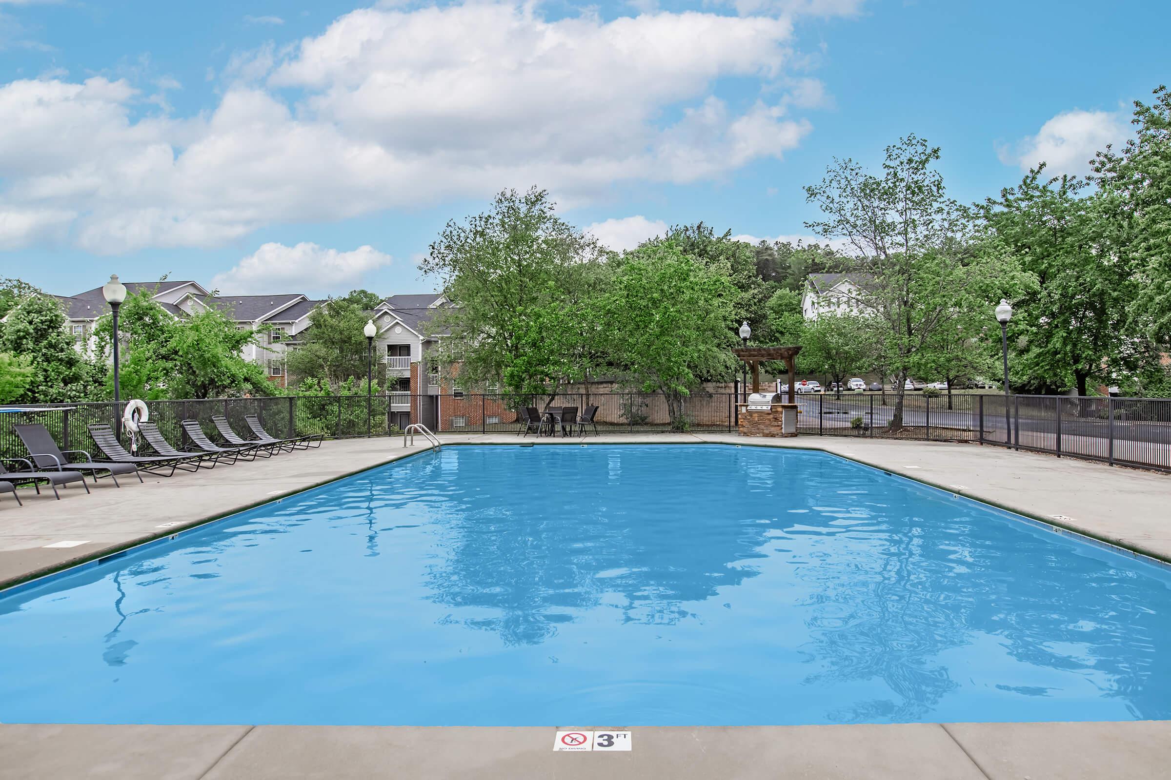 A spacious outdoor swimming pool surrounded by lounge chairs and trees. The clear blue water reflects the sky, with a shallow end marked at 3 feet. Nearby, there's a shaded area with a gazebo, and residential buildings in the background under a partly cloudy sky.