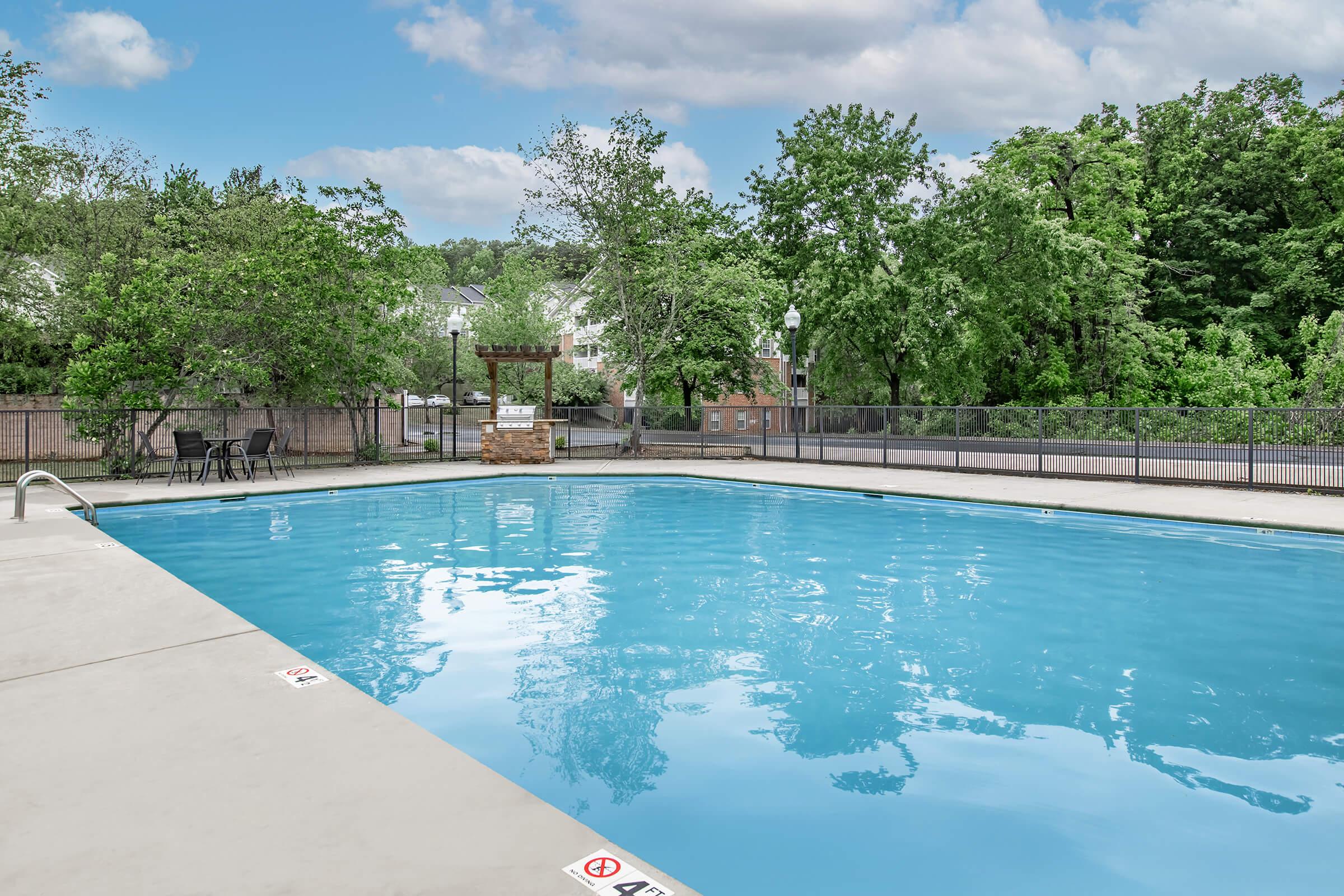 A clear blue swimming pool surrounded by a concrete deck and a black fence. Lush green trees and foliage are visible in the background, with a few chairs placed nearby. The scene conveys a peaceful outdoor atmosphere, ideal for relaxation.