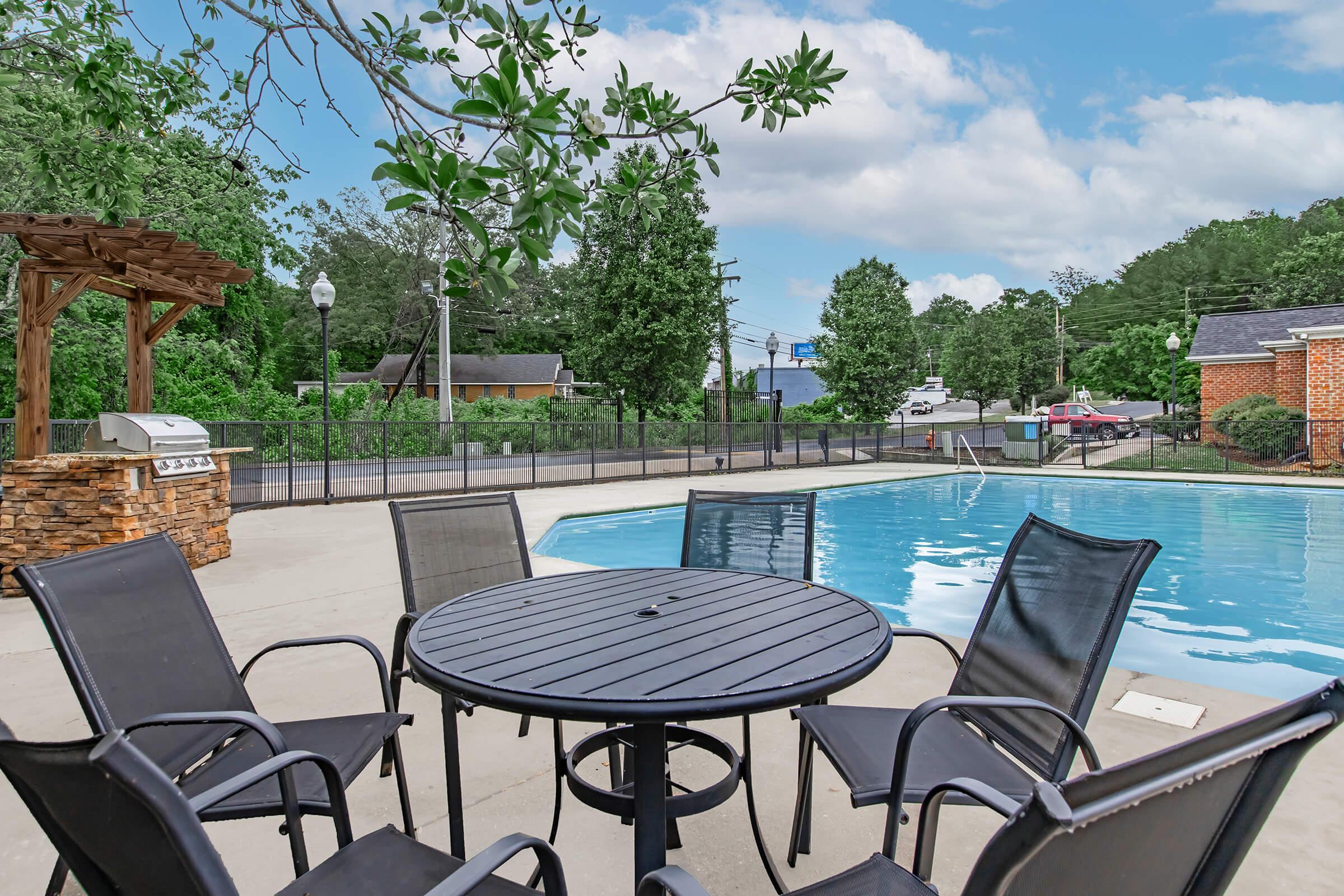 A poolside scene featuring a round black table surrounded by six chairs, with a grill nearby under a wooden structure. The pool is clear and inviting, set against a backdrop of trees and buildings under a partly cloudy sky.