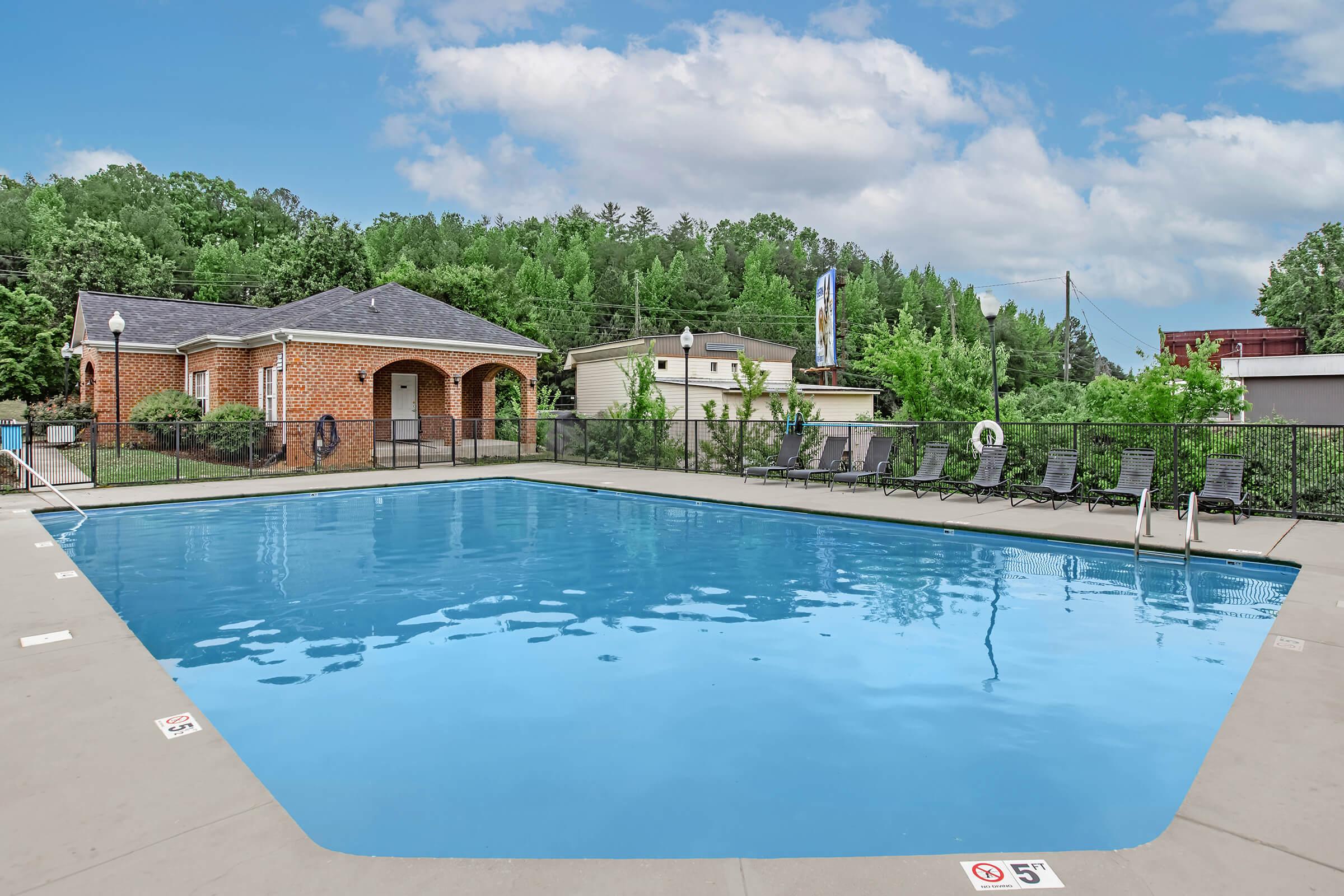 A clear blue swimming pool surrounded by a concrete deck, with several black loungers positioned along the side. In the background, there's a brick building with an arched entrance, surrounded by trees and greenery under a partly cloudy sky.