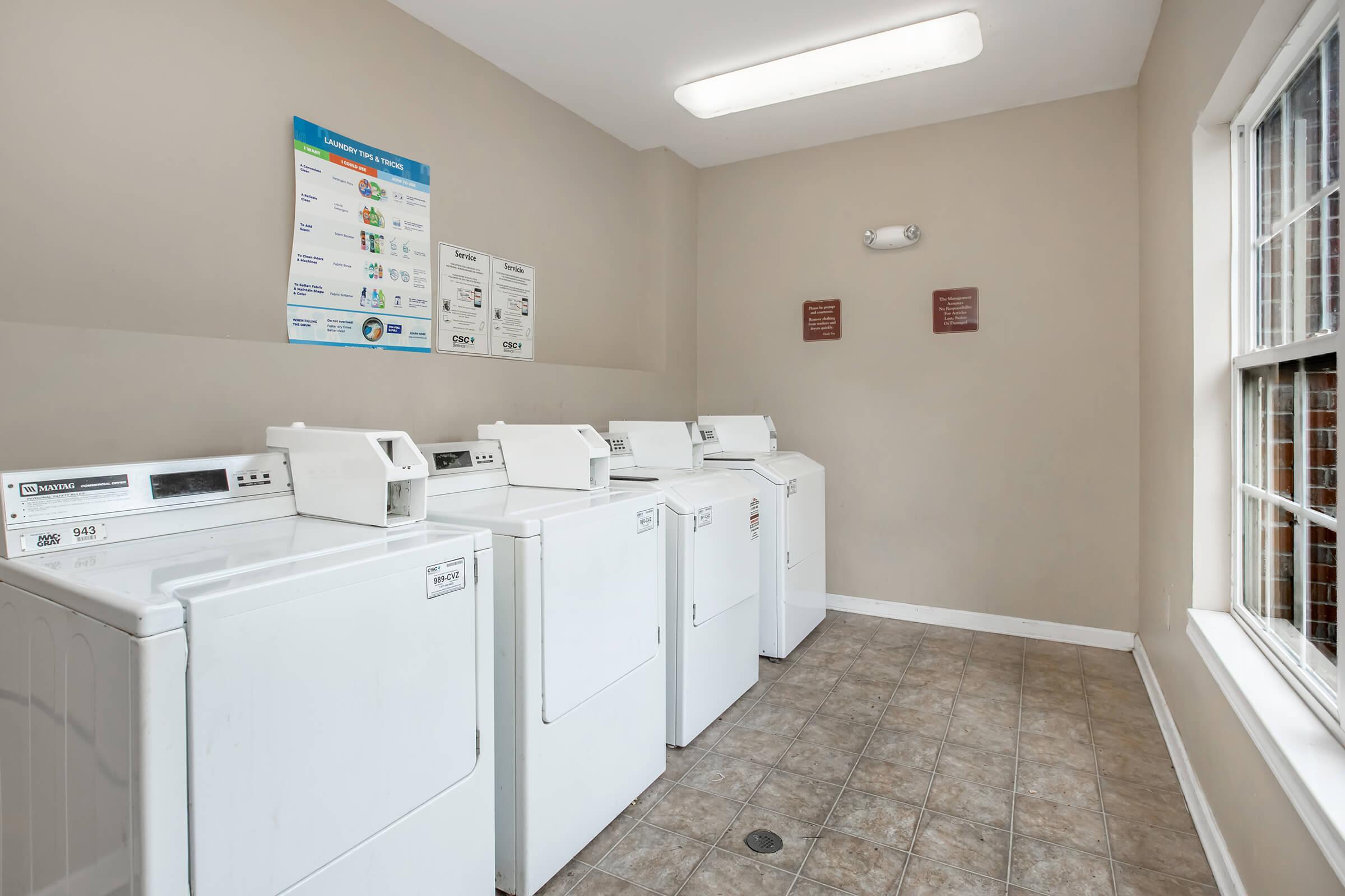 A clean laundry room featuring several white washing machines and dryers lined up against a wall. There is a large window providing natural light, along with a bulletin board and signs on the wall offering laundry instructions and guidelines. The floor is tiled for easy maintenance.