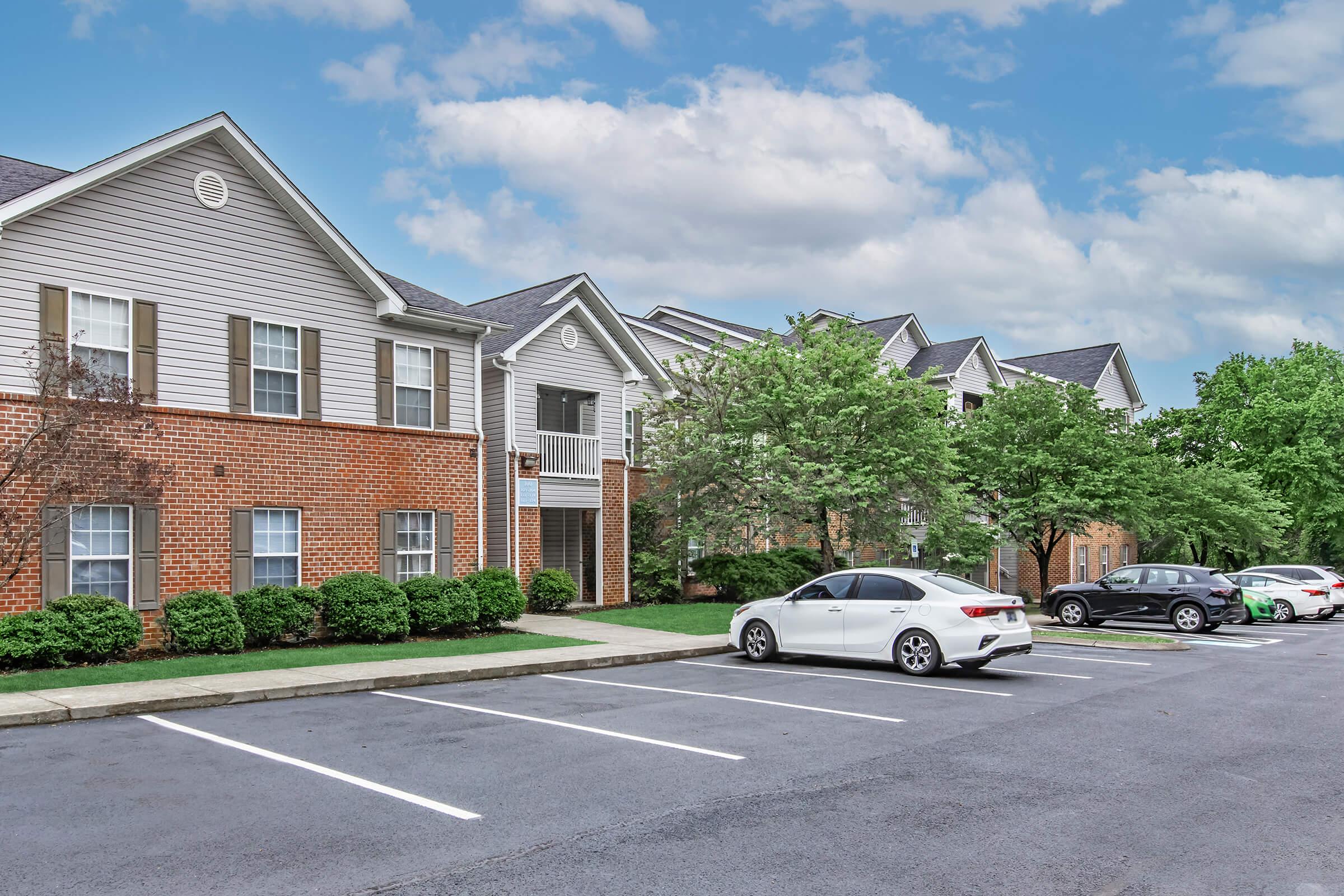 A view of a residential complex showcasing multiple two-story buildings with a mix of brick and siding exteriors. The scene includes green landscaping, trees, and a parking lot with several parked cars beneath a partly cloudy sky.
