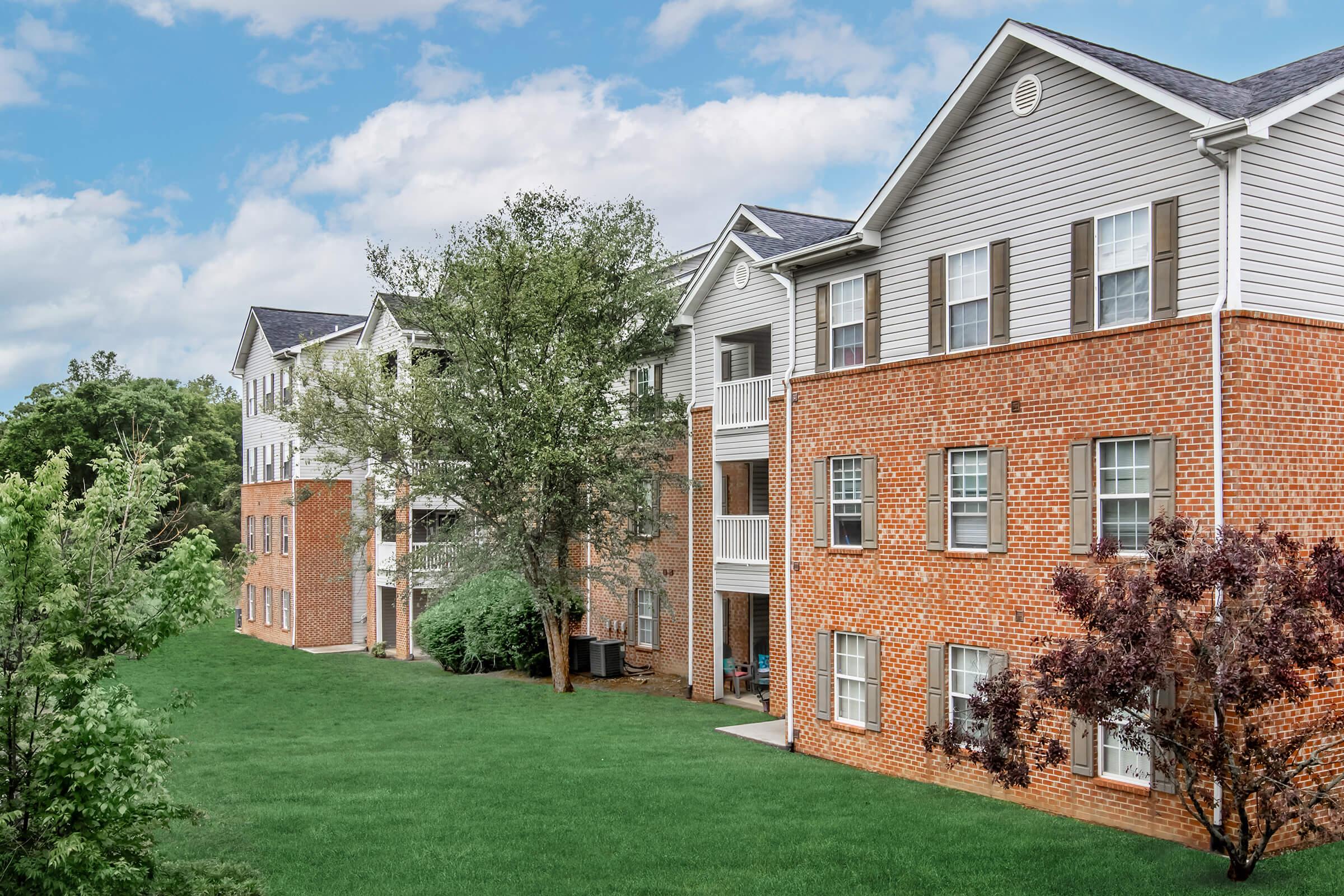 Apartment buildings with a mix of brick and siding, set against a grassy area and trees. The sky is partly cloudy, creating a serene atmosphere.