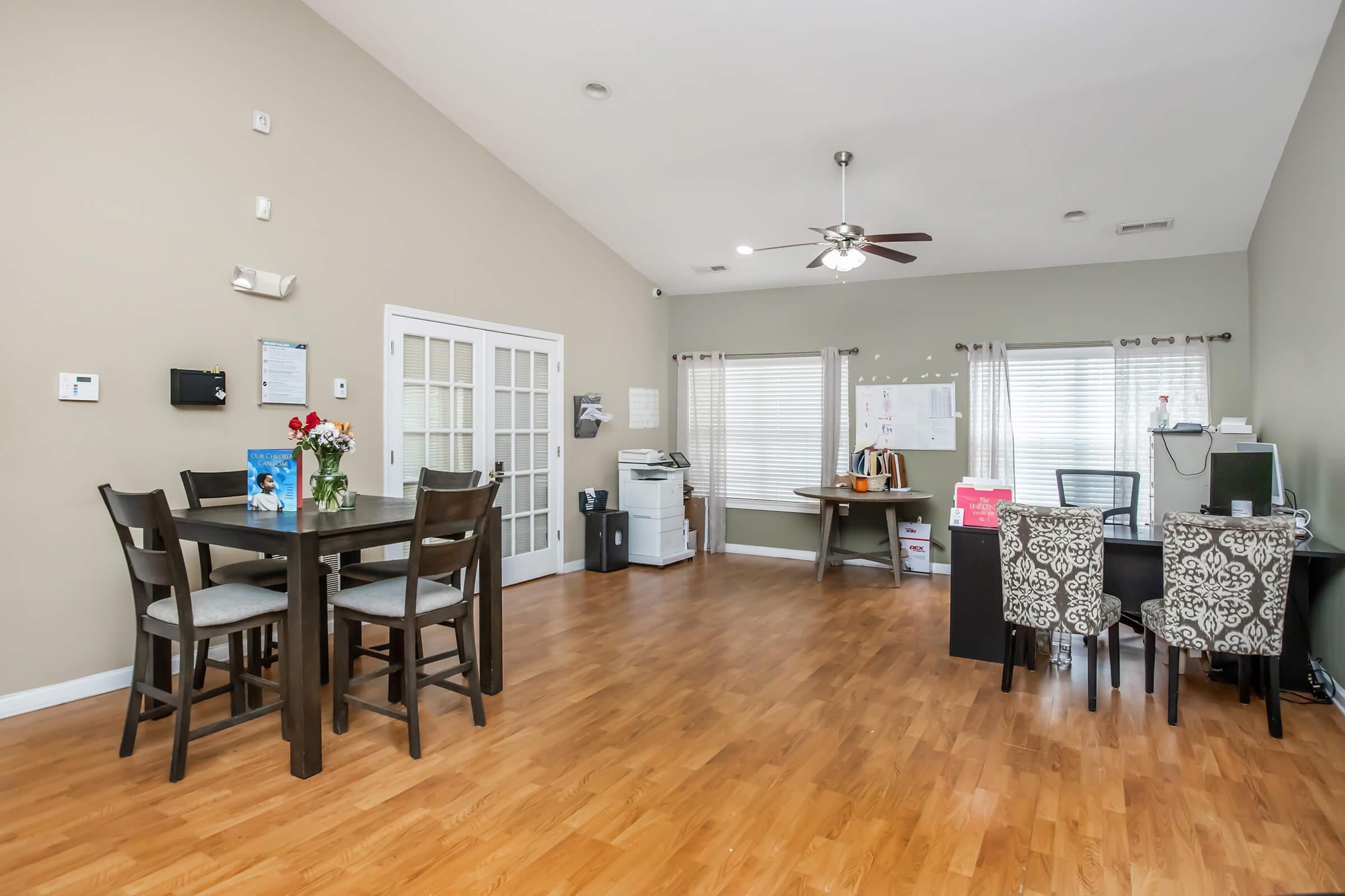 A spacious interior of a modern room featuring a dining table with four chairs, a work desk with a computer, and a printer. The walls are painted light gray, and there are large windows allowing natural light. A floral arrangement is on the table, and there are decorative chairs at the work desk.