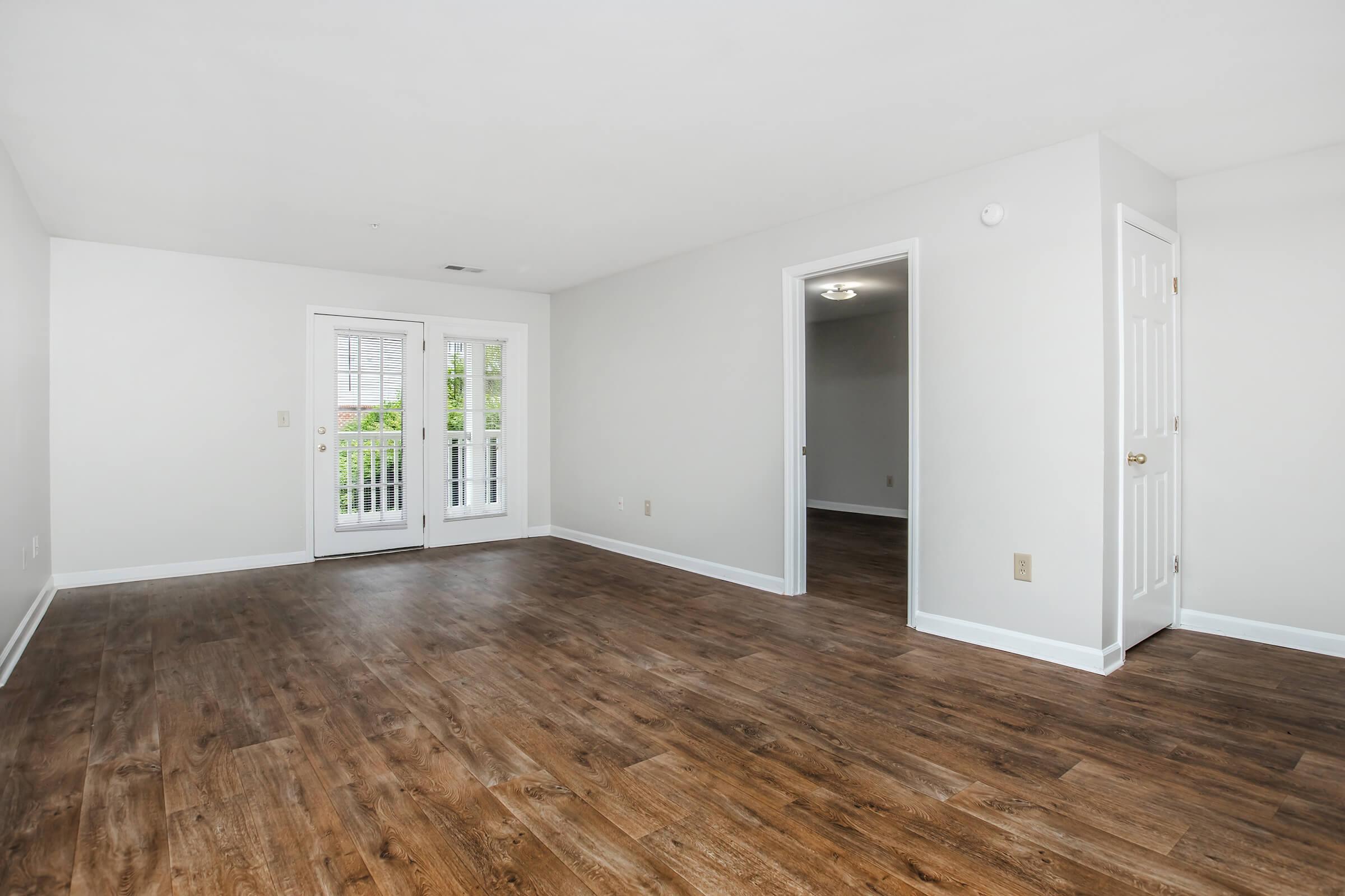 Empty room with light-colored walls and a hardwood floor. A set of double doors leads to a balcony, while a door on the right opens to another room. The space is well-lit and airy, showcasing a minimalist aesthetic with no furniture present.