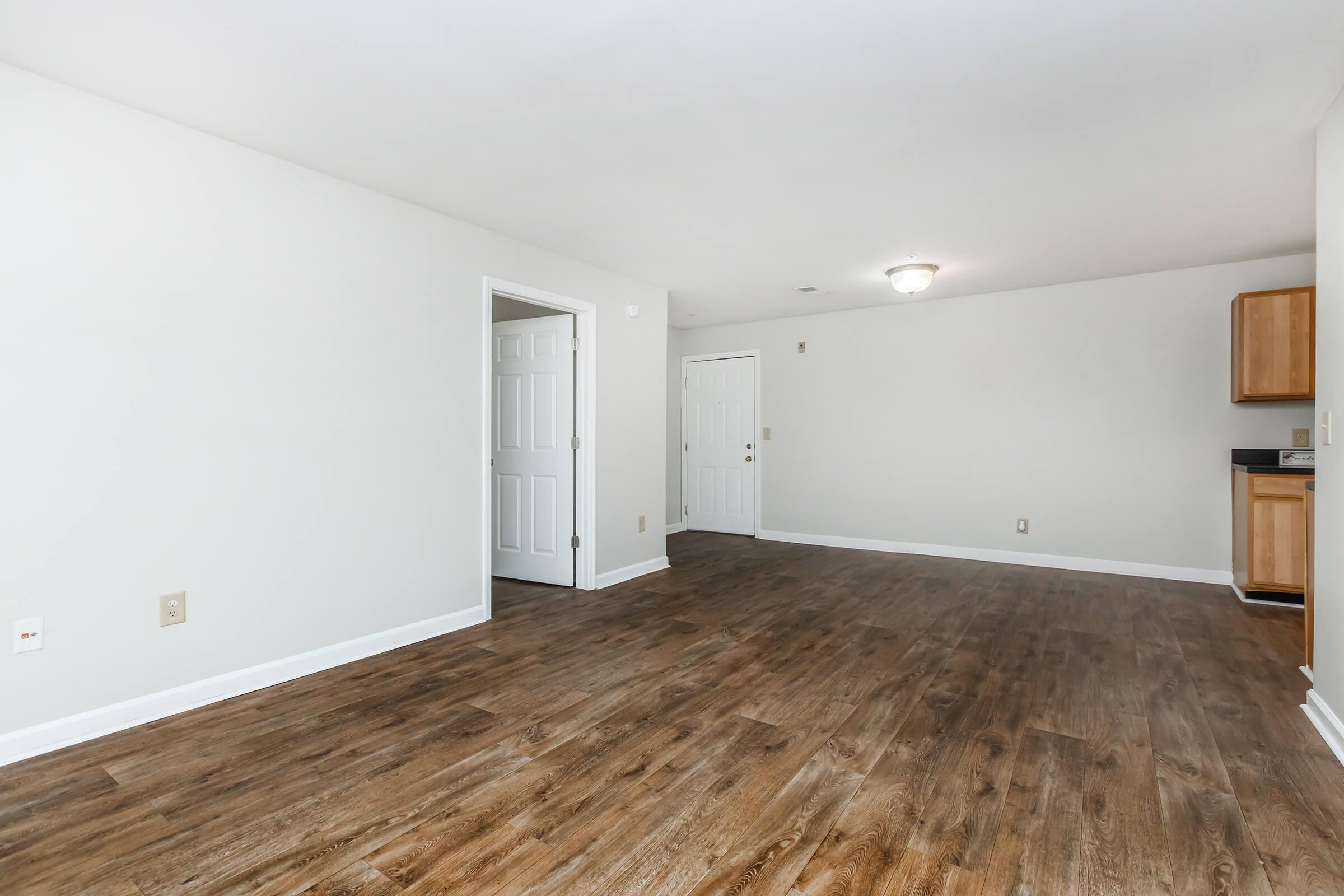 A spacious, well-lit room with light gray walls and wooden flooring. There is an open area with a doorway on the left leading to another room and a door on the right. The opposite wall features a kitchenette with wooden cabinets, creating a simple and modern living space.