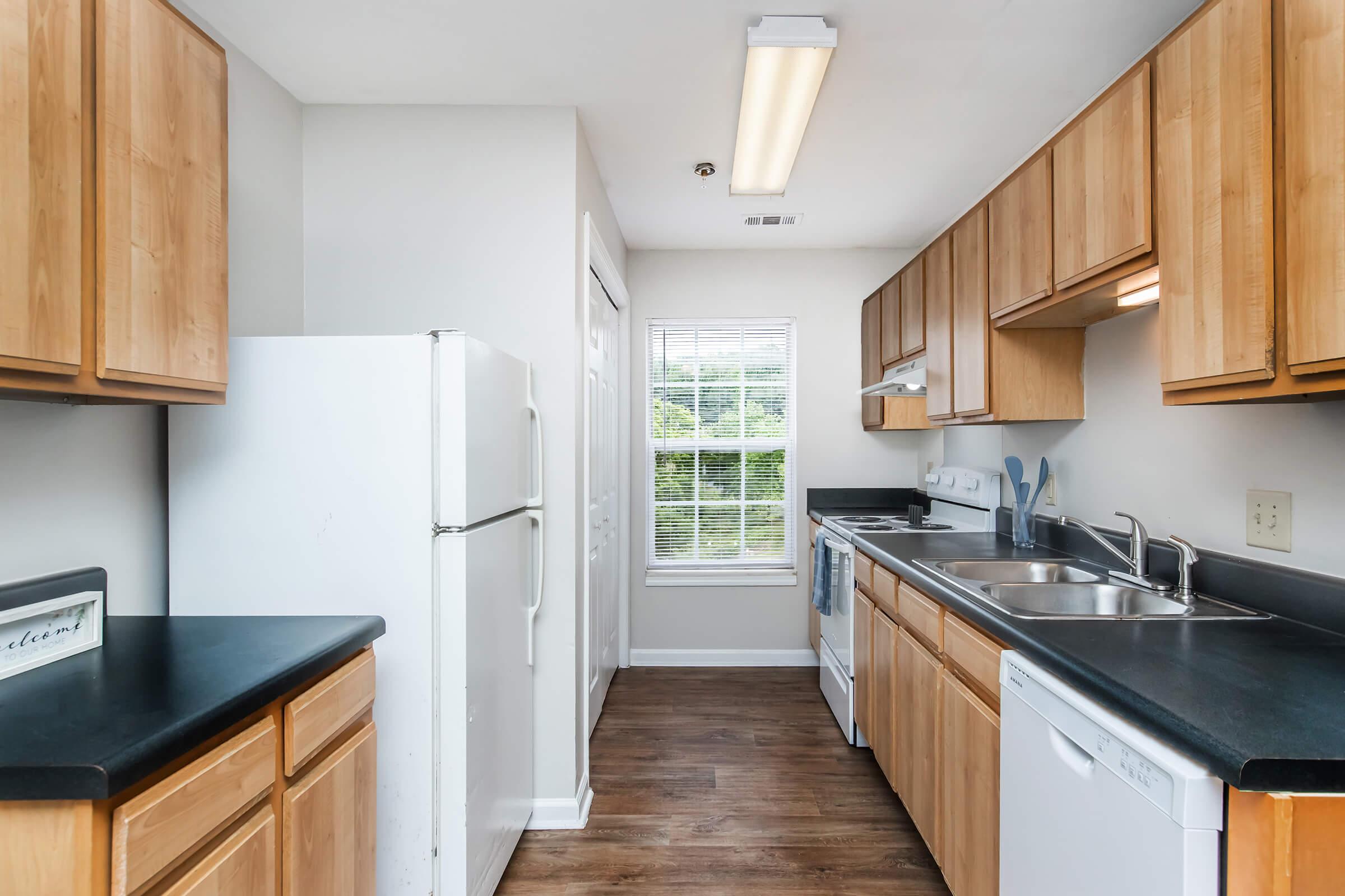 A modern kitchen featuring wooden cabinets, a white refrigerator, a stove, and a dishwasher. The countertops are dark, and there is a window allowing natural light in. The floor is wooden, creating a warm and inviting atmosphere.