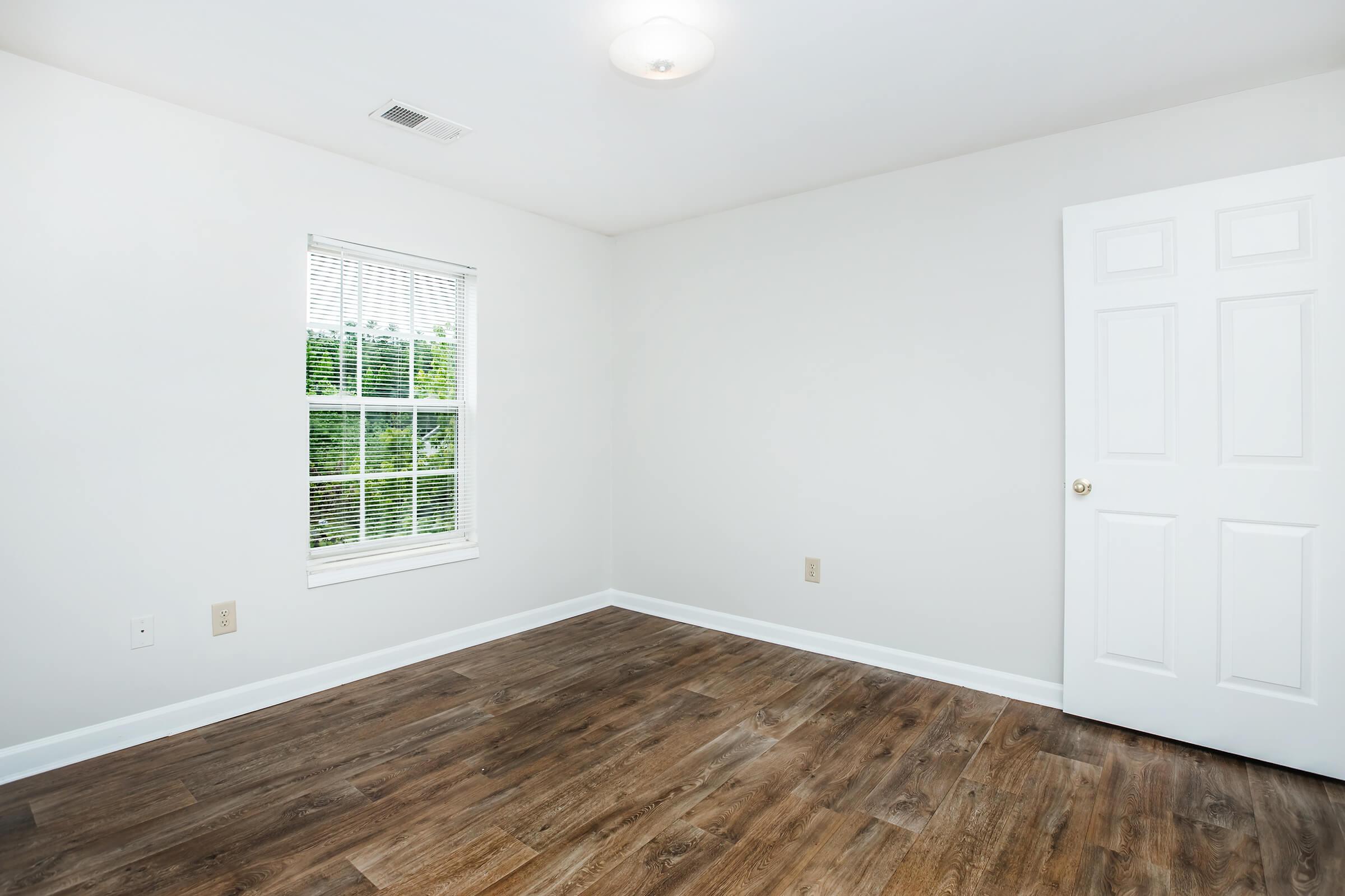 Empty room with light-colored walls and a wooden floor. A window with blinds allows natural light in, while a light fixture is mounted on the ceiling. There is a closed white door on the right side. The space is clean and uncluttered, suitable for various uses or furnishings.