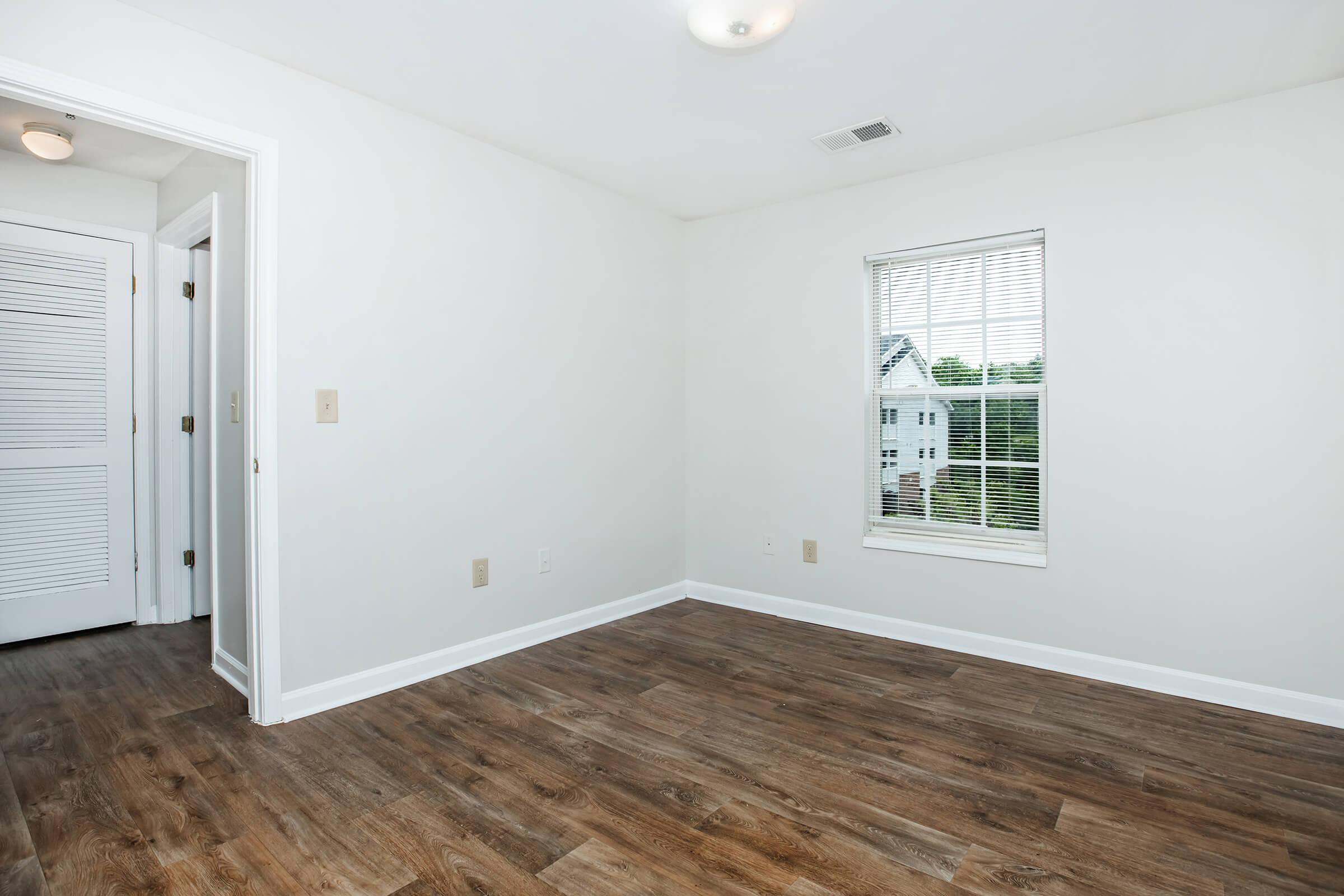 A bright, empty room featuring light-colored walls and a hardwood floor. A window with blinds allows natural light to enter, offering a view of the outside. There is a door leading to another space, and overall, the room appears clean and ready for furnishing.