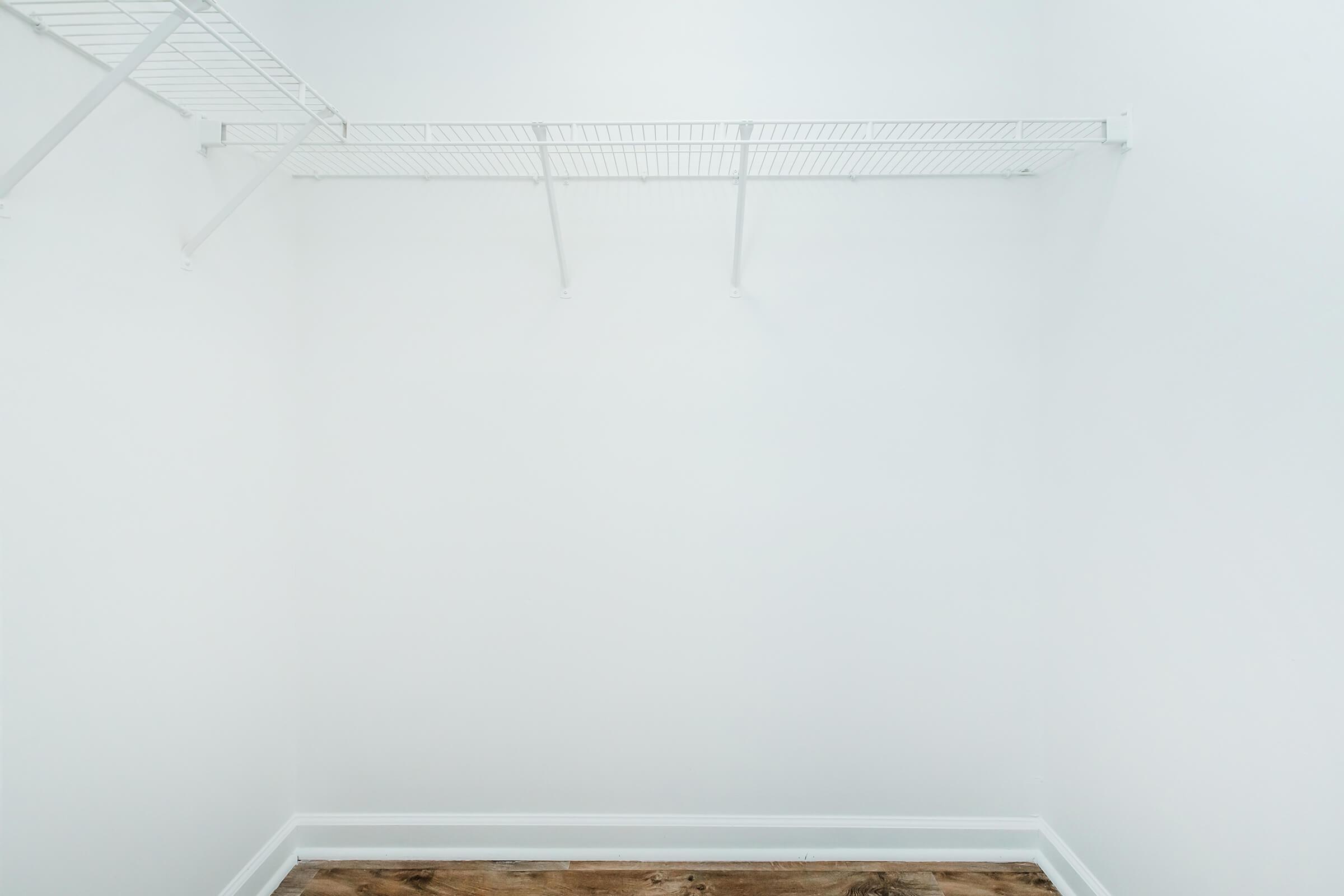A minimalist empty closet with white walls and a wooden floor. The closet features two wire shelves on the left side and one in the center, providing a clean and open space for storage.