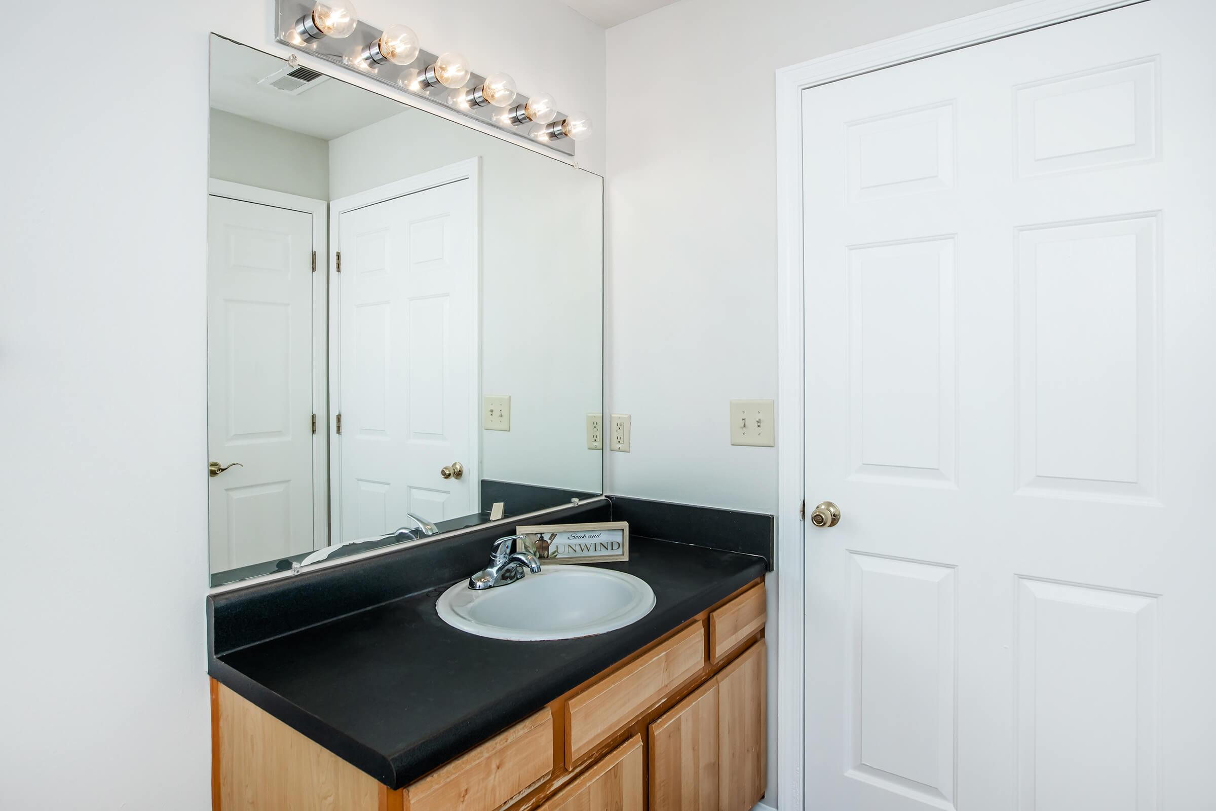 A bathroom featuring a black countertop with a sink, a large mirror above, and a light fixture with multiple bulbs. The cabinetry is wooden, and there is a closed white door on the right side of the image. The walls are painted white, creating a clean and modern look.