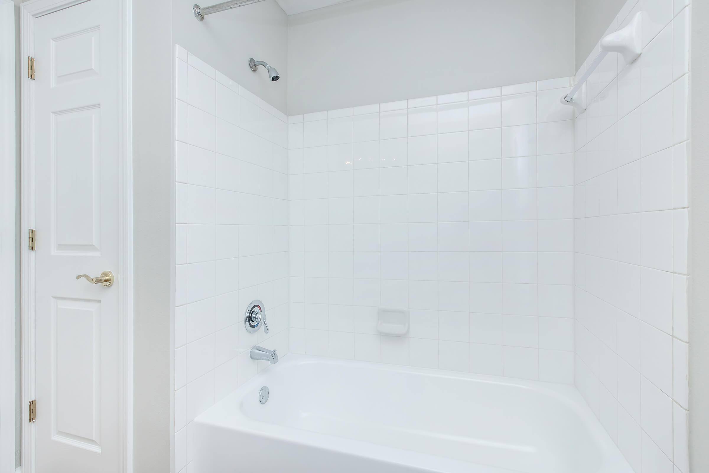 A clean, modern bathroom featuring a white tiled shower and bathtub. The space includes a showerhead, tub faucet, and a towel rack, with a light-colored wall and a white door visible in the frame. The overall look is bright and minimalistic.