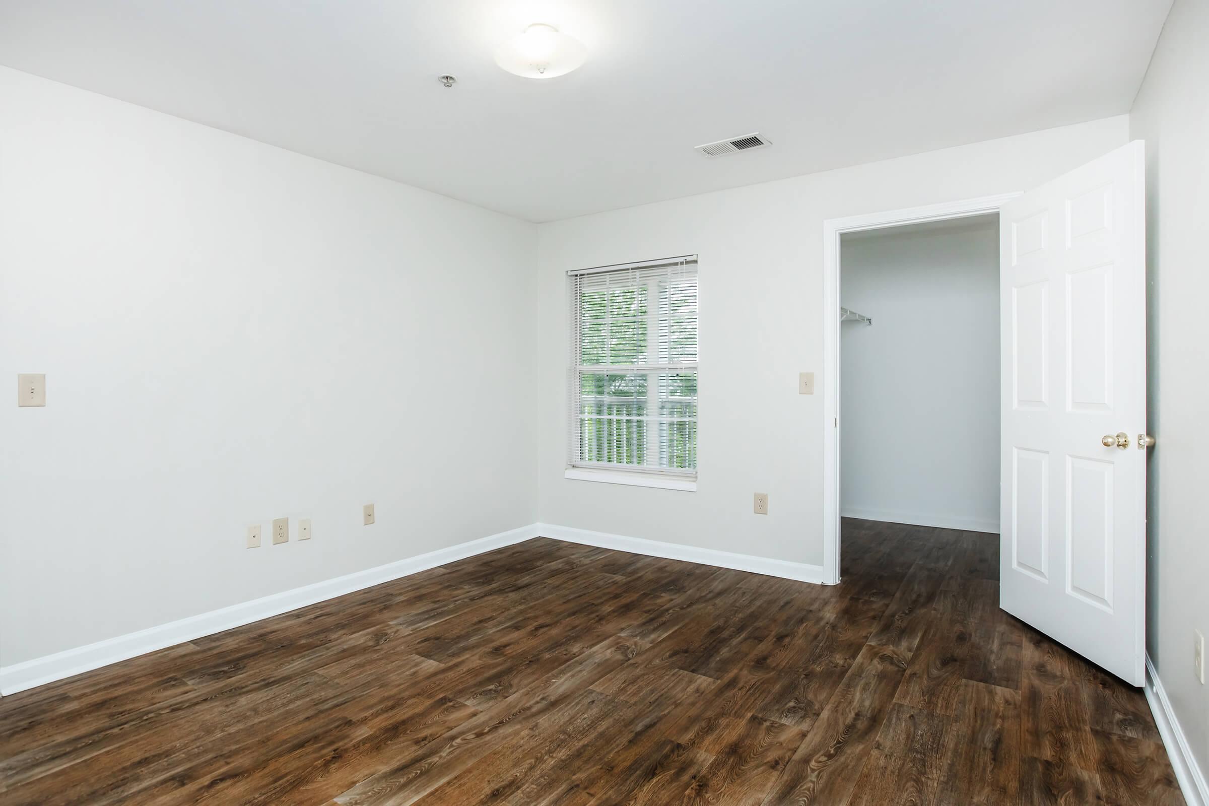 Empty room with light-colored walls, a window with blinds, and a wooden floor. A closet is visible through an open door on the right side. Natural light fills the space, creating a bright and airy atmosphere.