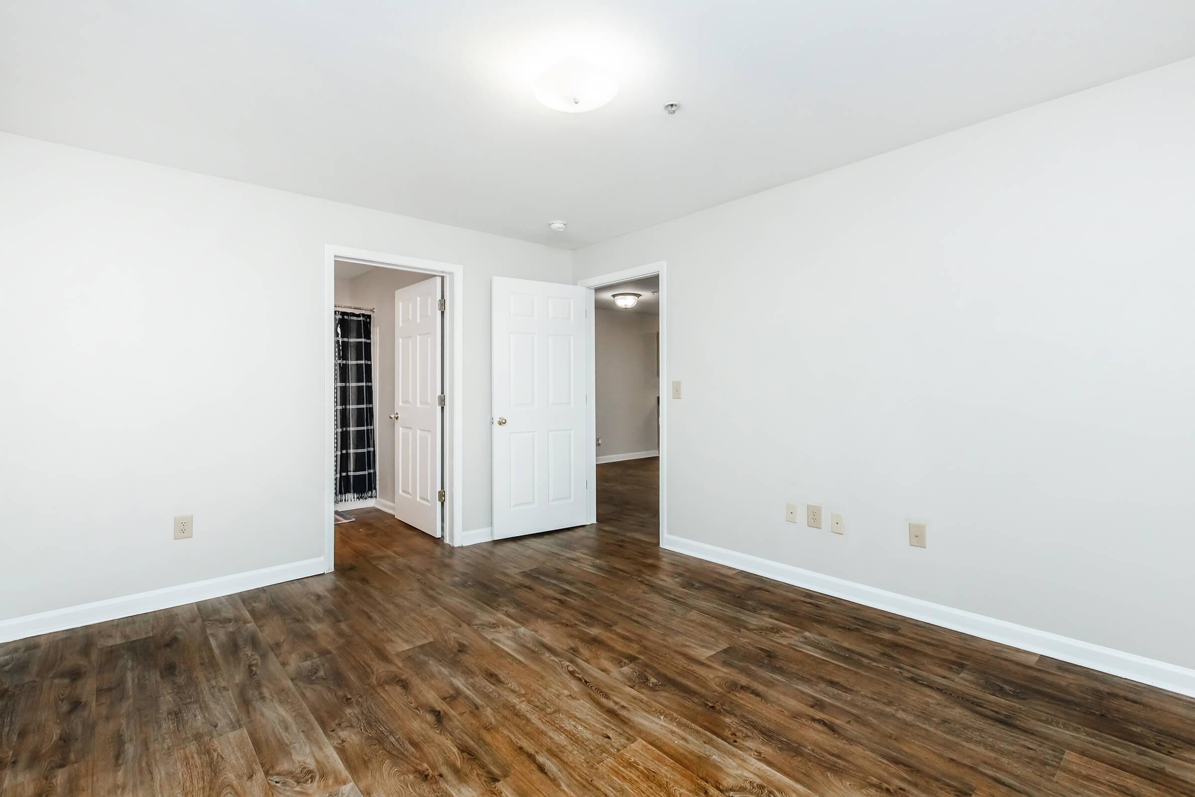 A spacious, empty room with light-colored walls and a wood-like floor. There are two open doors leading to other areas, and a small bathroom visible through one of the doors. The ceiling has a simple light fixture. The overall atmosphere is bright and clean, suitable for various uses.
