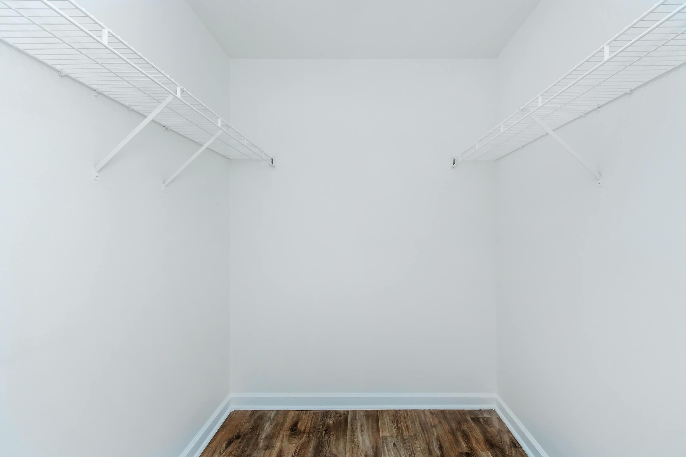 An empty closet with white walls and two wire shelves on opposite sides. The floor is made of wooden planks, and there is a sense of spaciousness in the room, emphasizing its simplicity and minimalism.