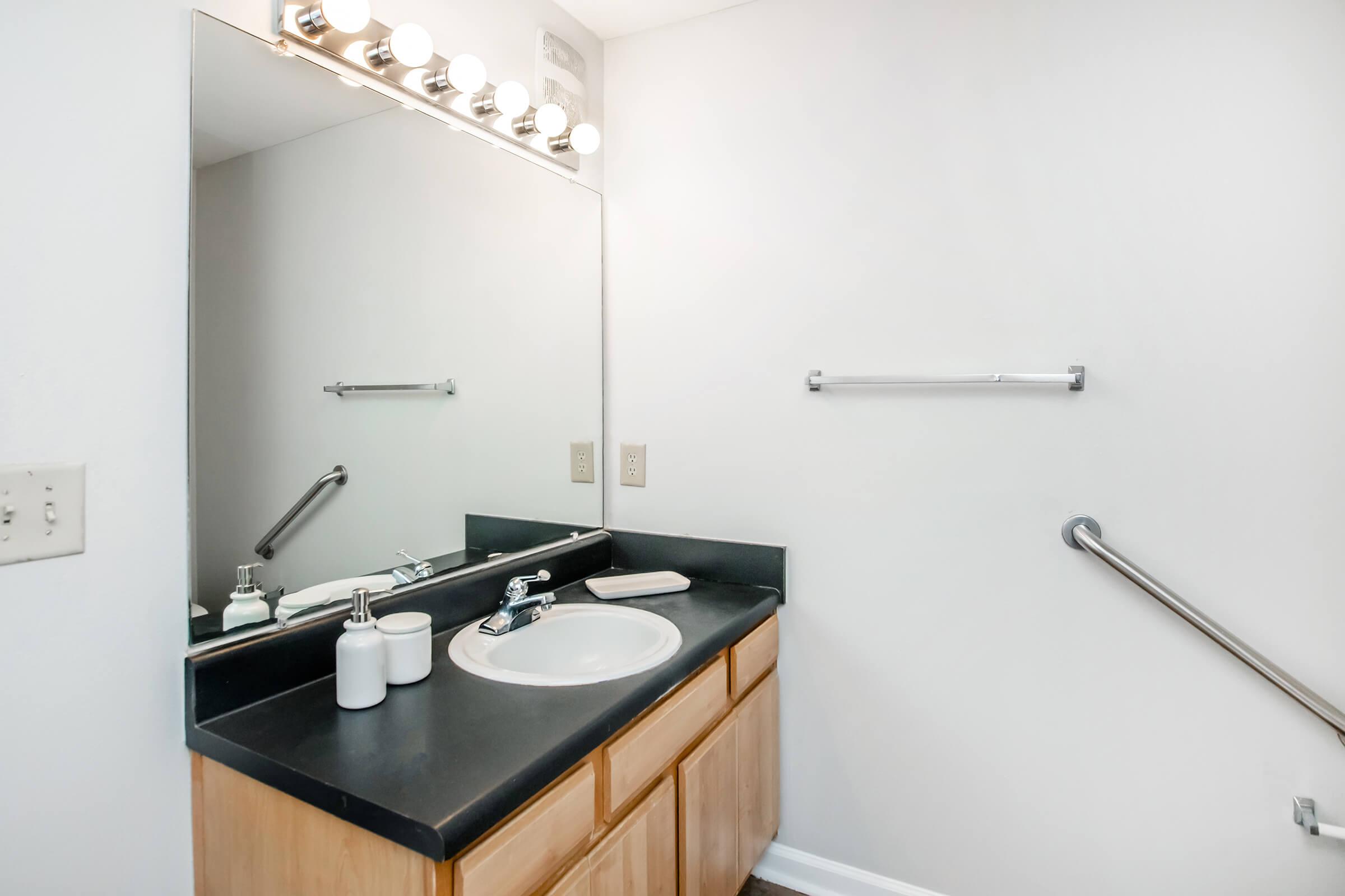 A clean, modern bathroom featuring a dark countertop with a sink, a wooden vanity with drawers, and a large mirror. Above the sink, there are light fixtures with multiple bulbs. The walls are painted white, and there is a towel bar and grab bar installed for accessibility.