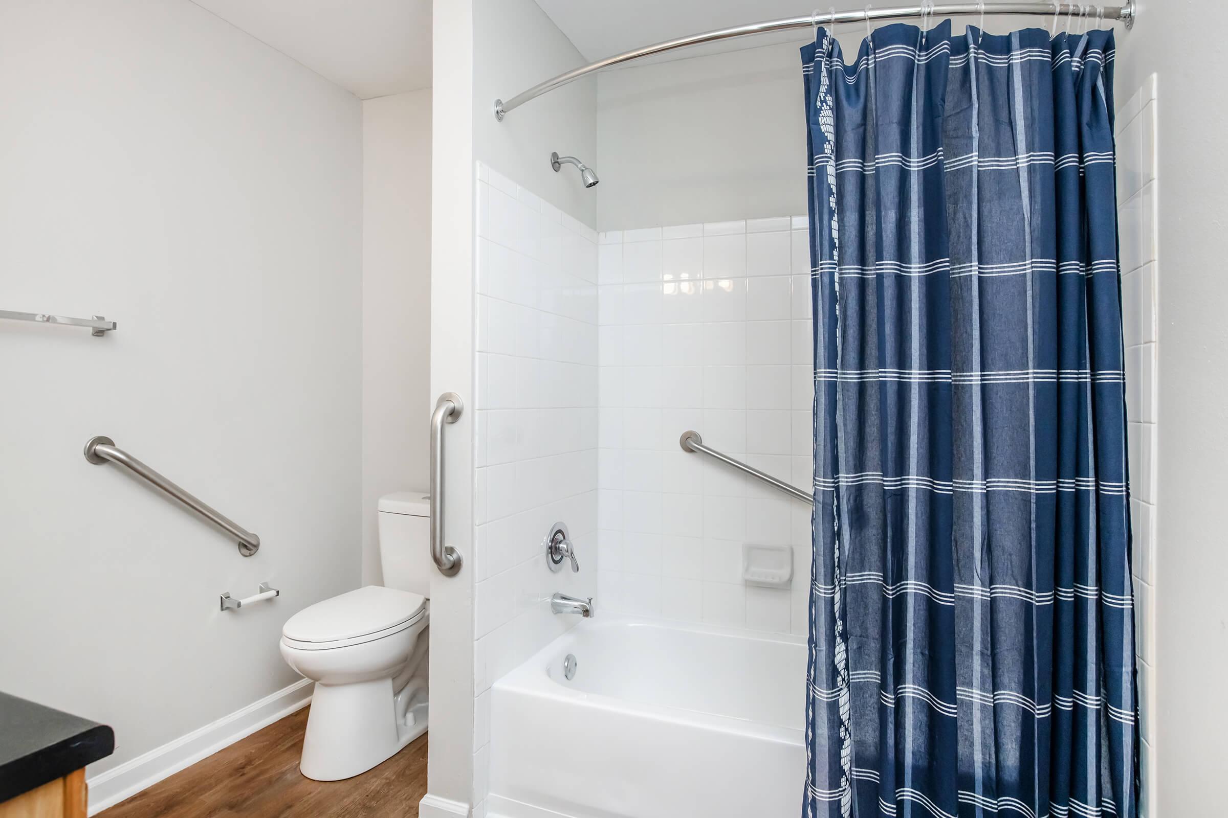 A modern bathroom featuring a white bathtub with a curved shower rod and navy blue checked shower curtain. There is a white toilet nearby, and grab bars are installed on the walls for accessibility. The floor is a light wood finish, complementing the neutral wall color.