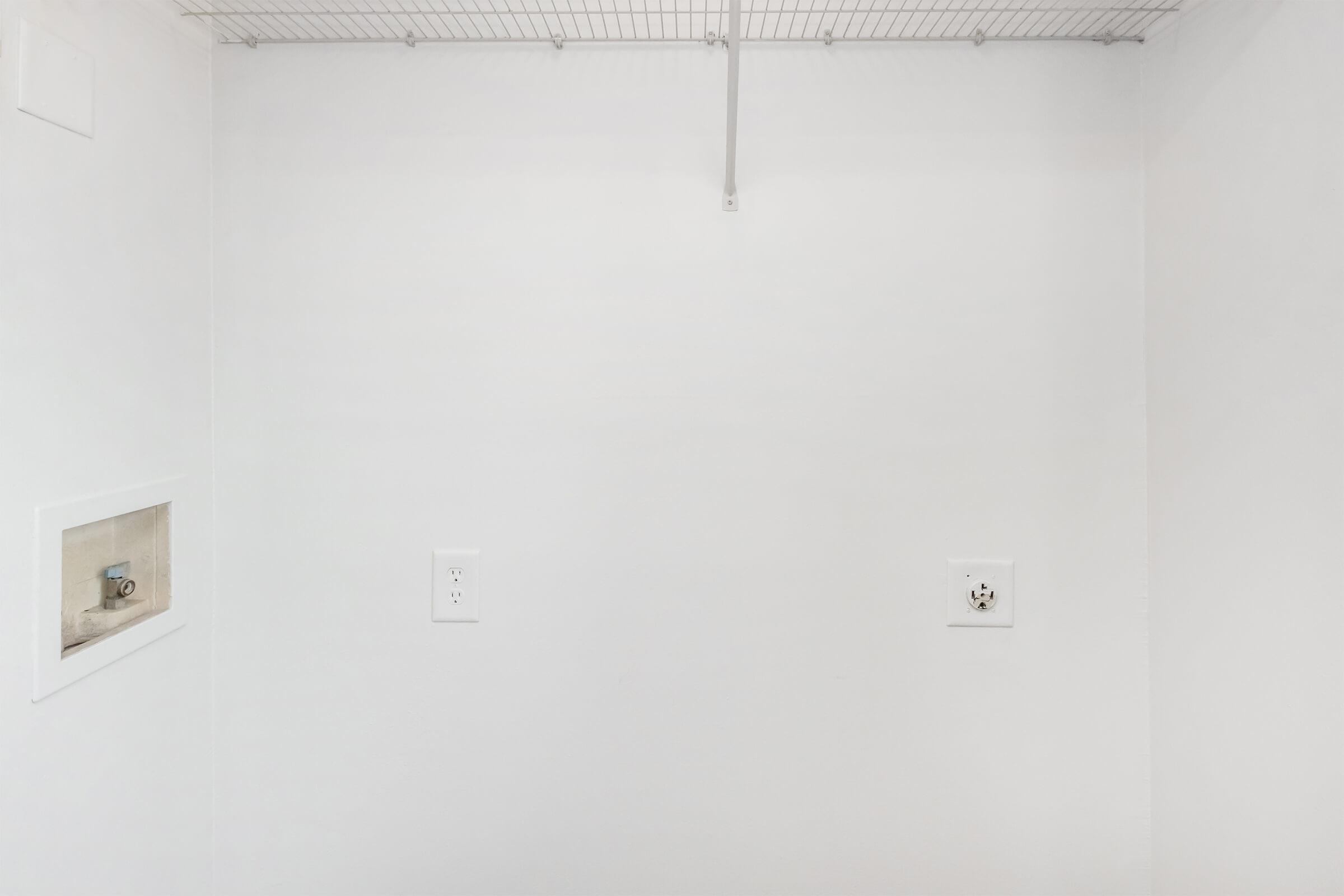 Empty closet space with white walls, featuring a wire shelf at the top. Two electrical outlets are visible on the left and right sides of the wall. The overall appearance is clean and minimalistic.