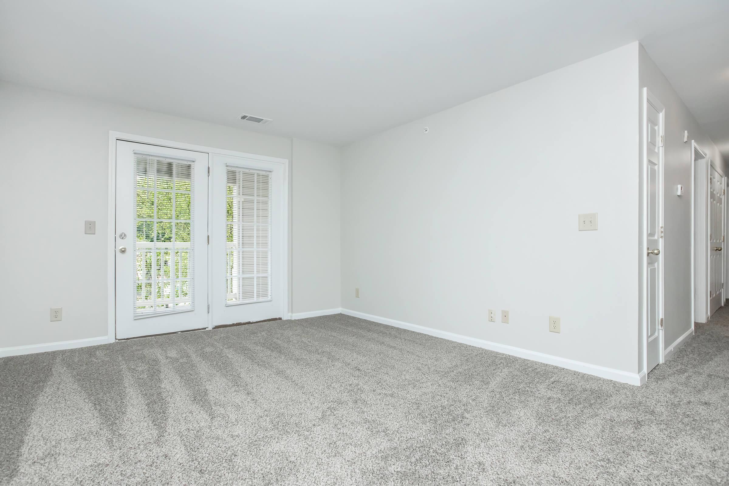 Empty room with light-colored walls and a carpeted floor, featuring two sliding glass doors leading to a balcony, and doorways on the right side leading to other rooms. Natural light is coming in, creating a bright and airy atmosphere. The space is clean and ready for furniture.