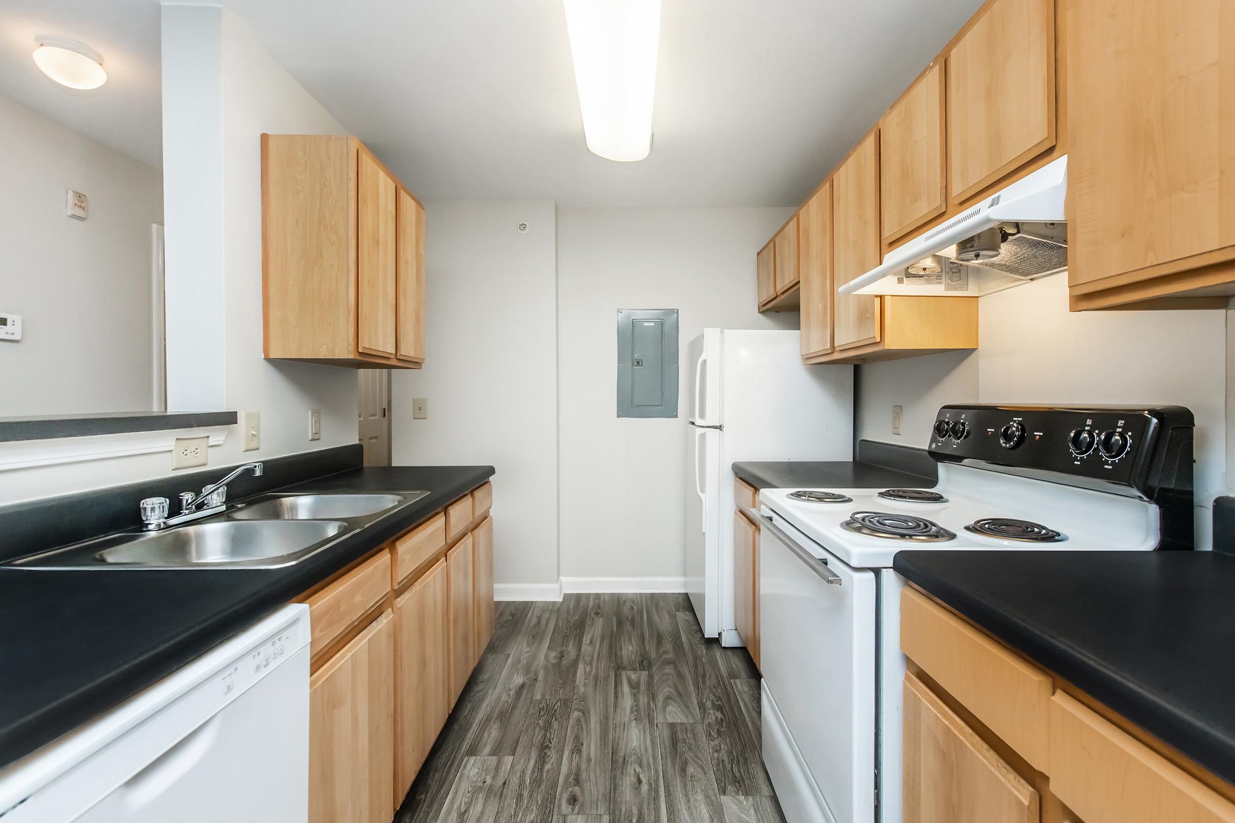 A modern kitchen featuring light wood cabinets, a black countertop, and stainless steel appliances. There are dual sinks, a dishwasher, a stove with an oven, and a refrigerator. The flooring is a dark laminate, and the walls are painted light gray, creating a clean and contemporary look.