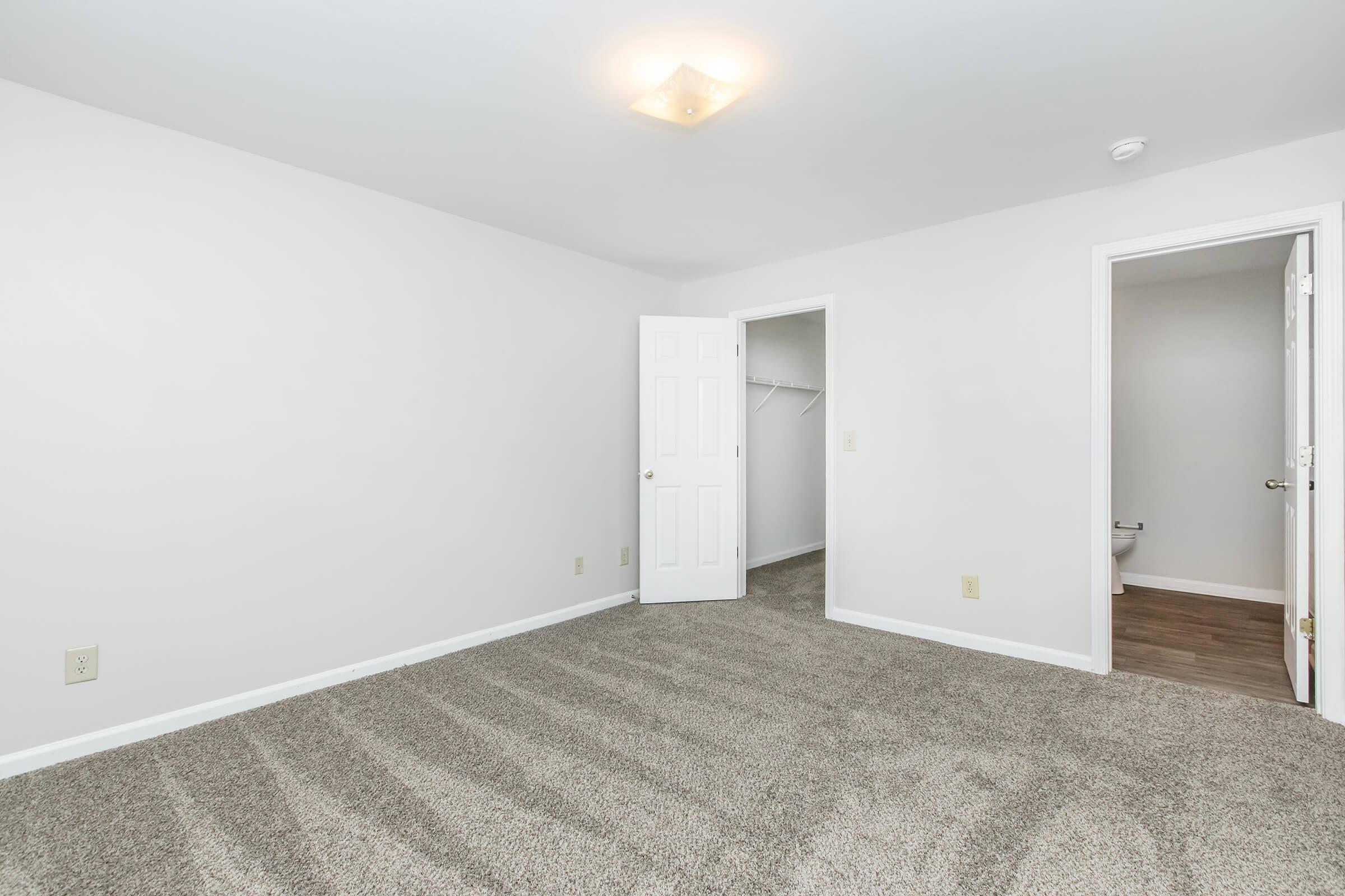 A well-lit, empty room with light gray walls and a carpeted floor. There are two doors: one leading to a closet and the other to a bathroom. The room has a simple ceiling light fixture and appears to be freshly painted and clean, creating a spacious and welcoming atmosphere.