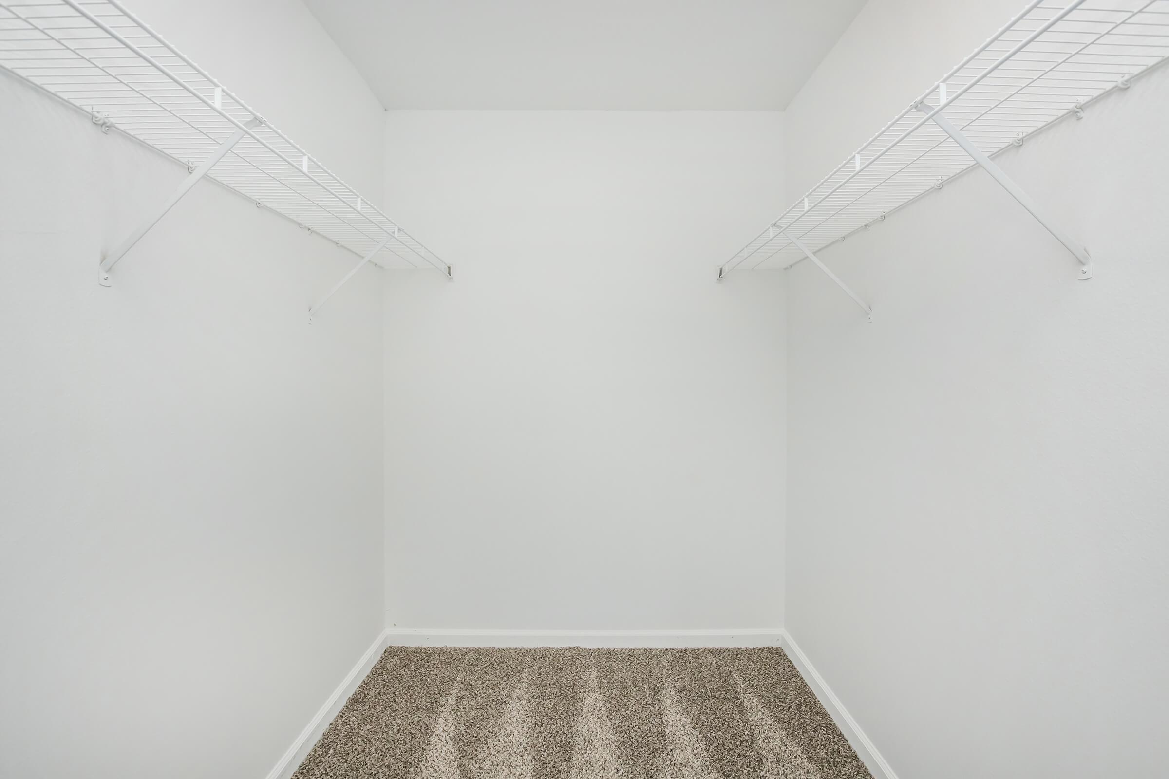 Empty closet with light gray walls and two wire shelves on opposite sides. The floor is covered with a soft, beige carpet, creating a clean and spacious appearance.