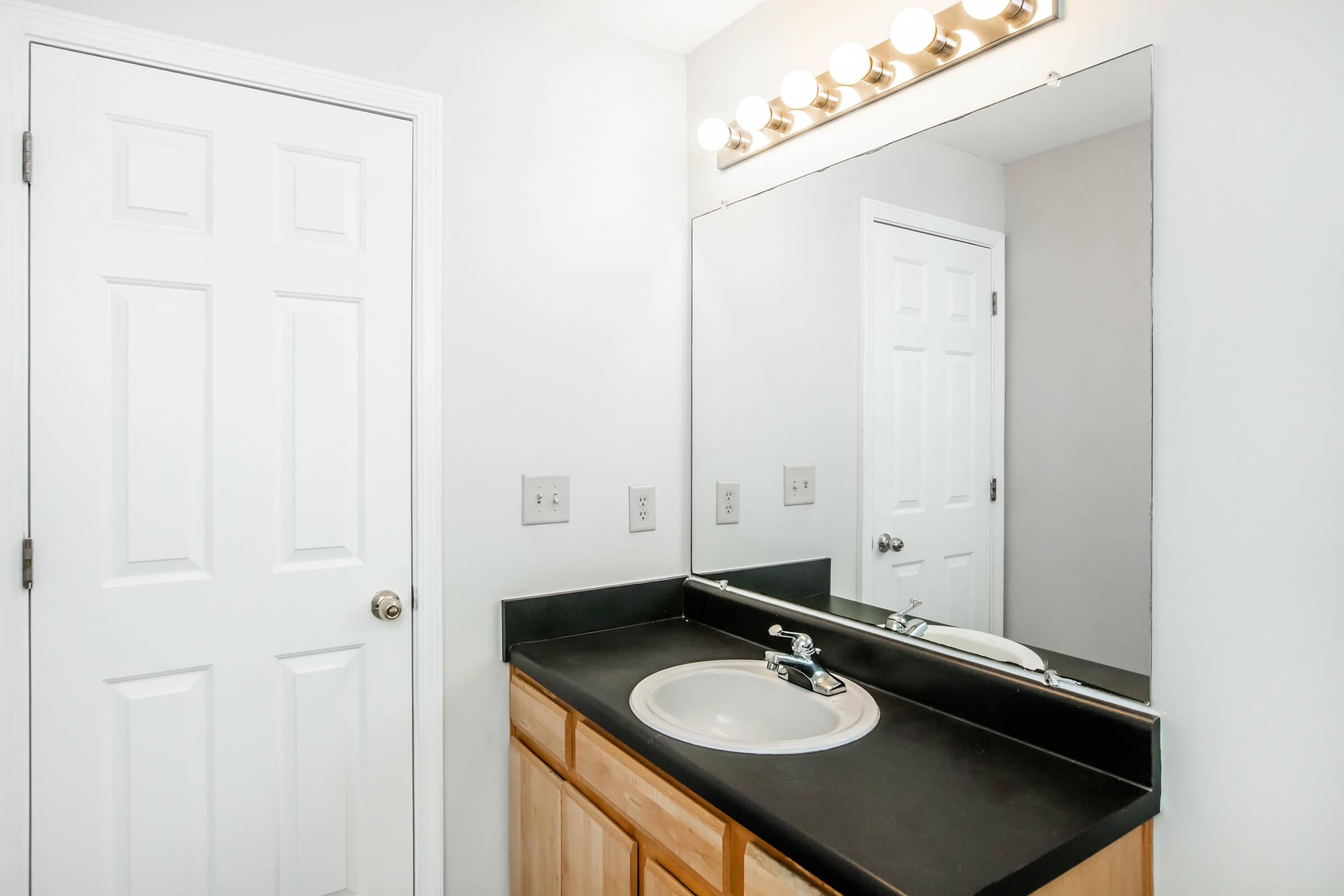 A modern bathroom featuring a black countertop with a sink and faucet, a large mirror above the sink, and a light fixture with multiple bulbs. The walls are painted white, and there is a closed white door in the background, adding to the clean, minimalist design.