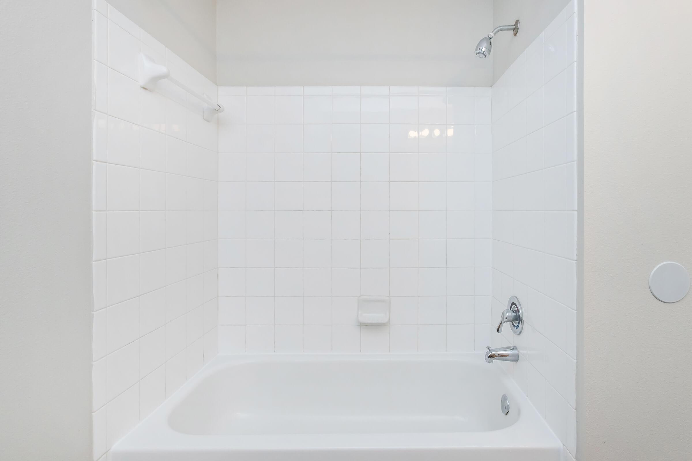 A clean, empty bathroom with a white tiled shower area and a bathtub. The walls are light-colored, and there are no personal items or decorations present. A showerhead is mounted on the wall, and there is a towel rack beside it. The overall appearance is modern and minimalistic.