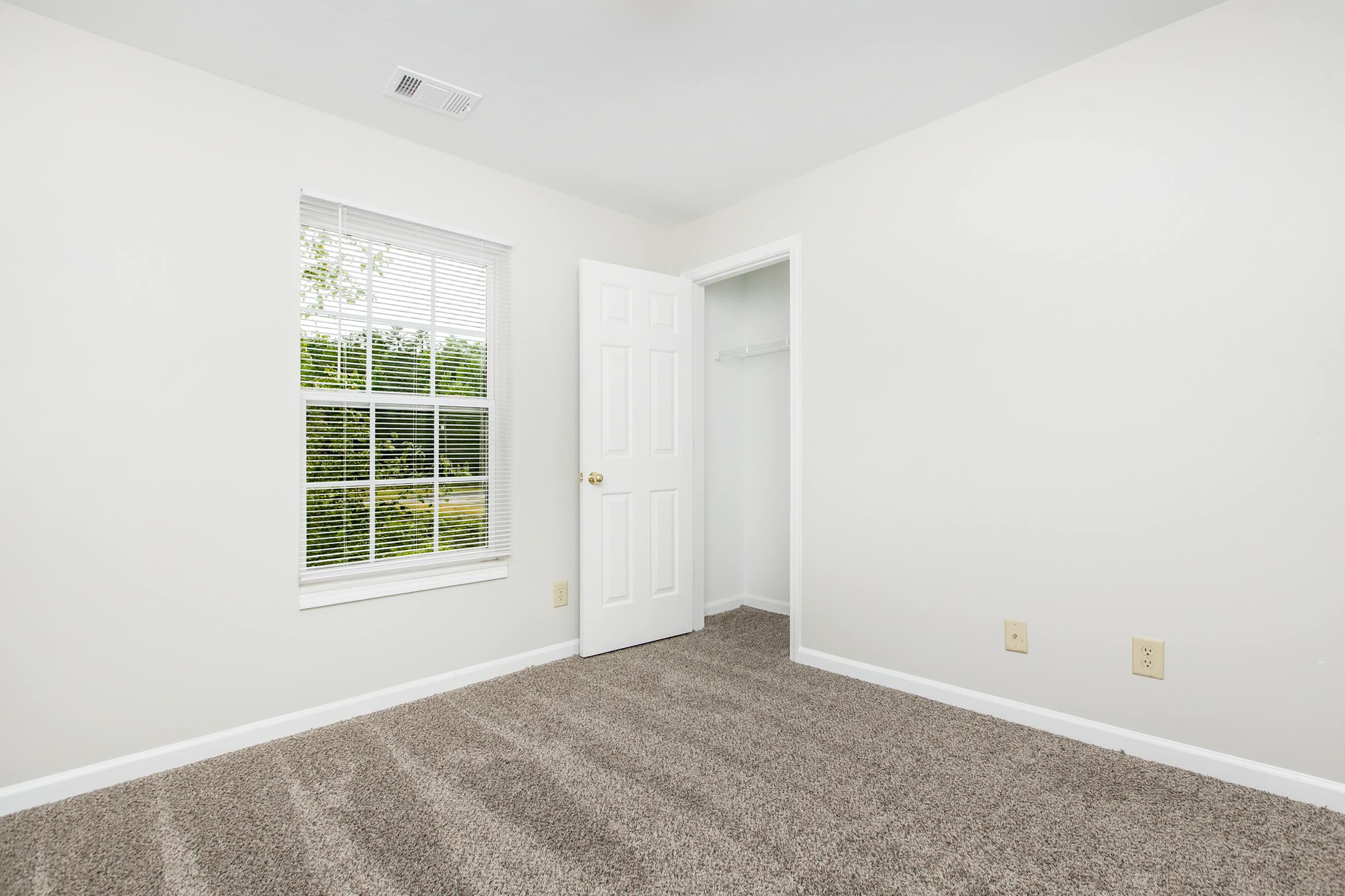 A vacant room with light-colored walls, a window with blinds allowing natural light, and a closet with a door ajar. The floor is covered in beige carpeting, creating a clean and spacious appearance.