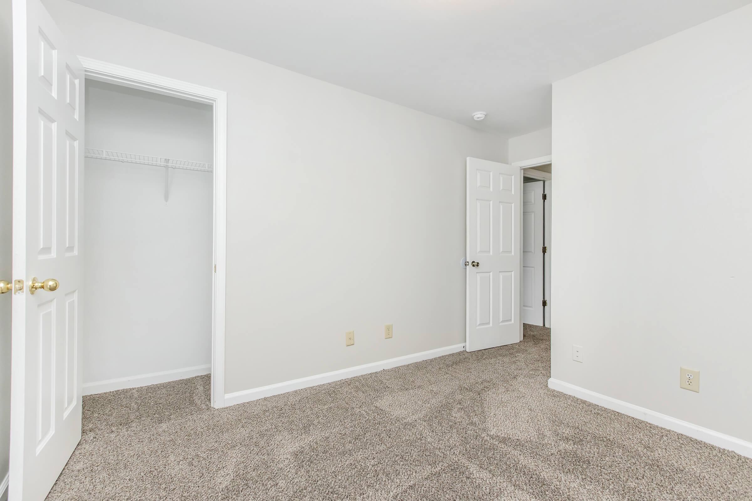 A spacious, empty room with light gray walls and beige carpet. There are two white doors: one leads to a small closet and the other opens to another part of the room. The room is well-lit and has a clean, minimalistic appearance, making it suitable for various uses.