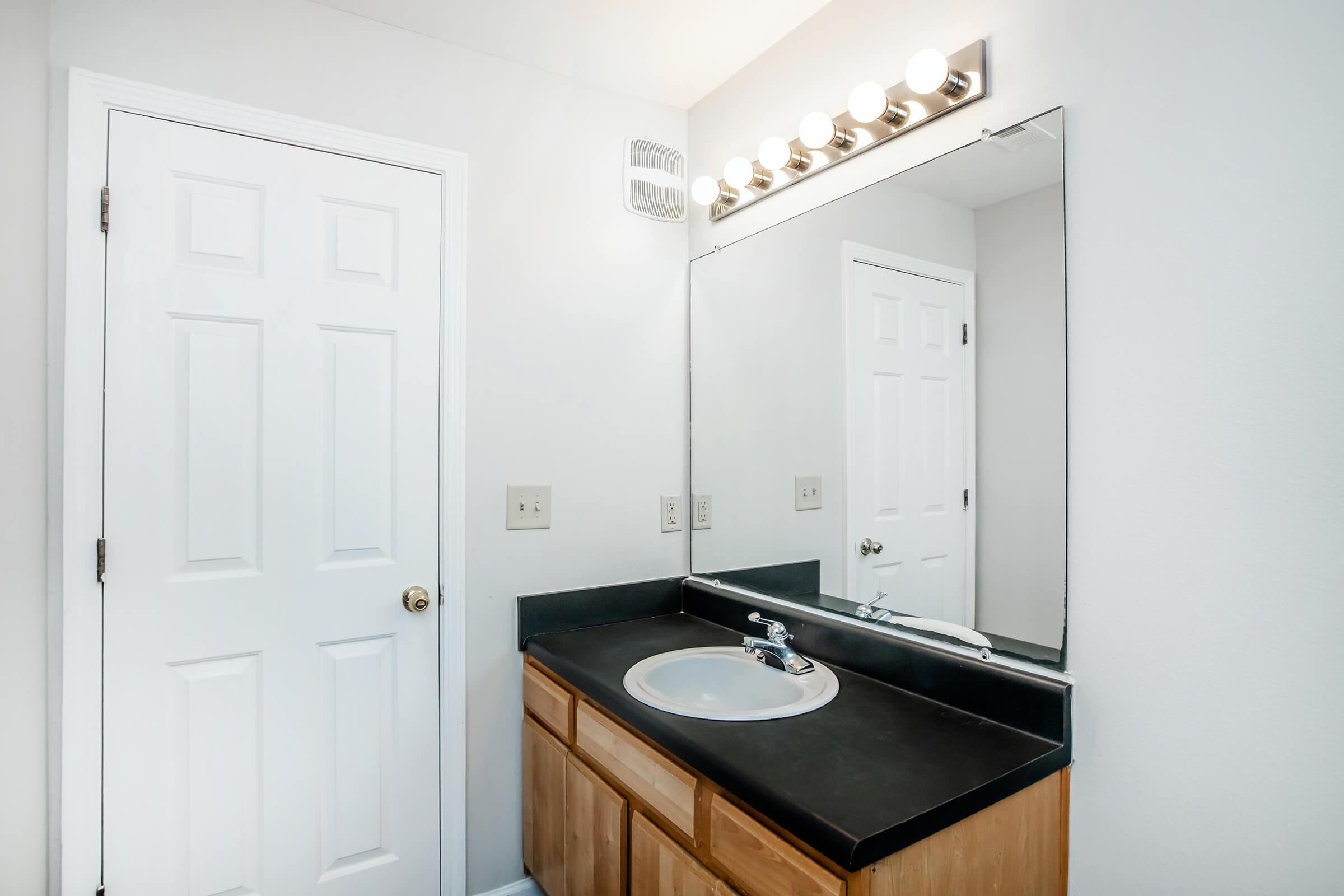 A clean, modern bathroom featuring a simple vanity with a sink, a large mirror above, and a lighted vanity fixture. The walls are painted white, and there is a door leading to another room. The countertop is dark, contrasting with the light-colored cabinetry. The overall feel is bright and spacious.
