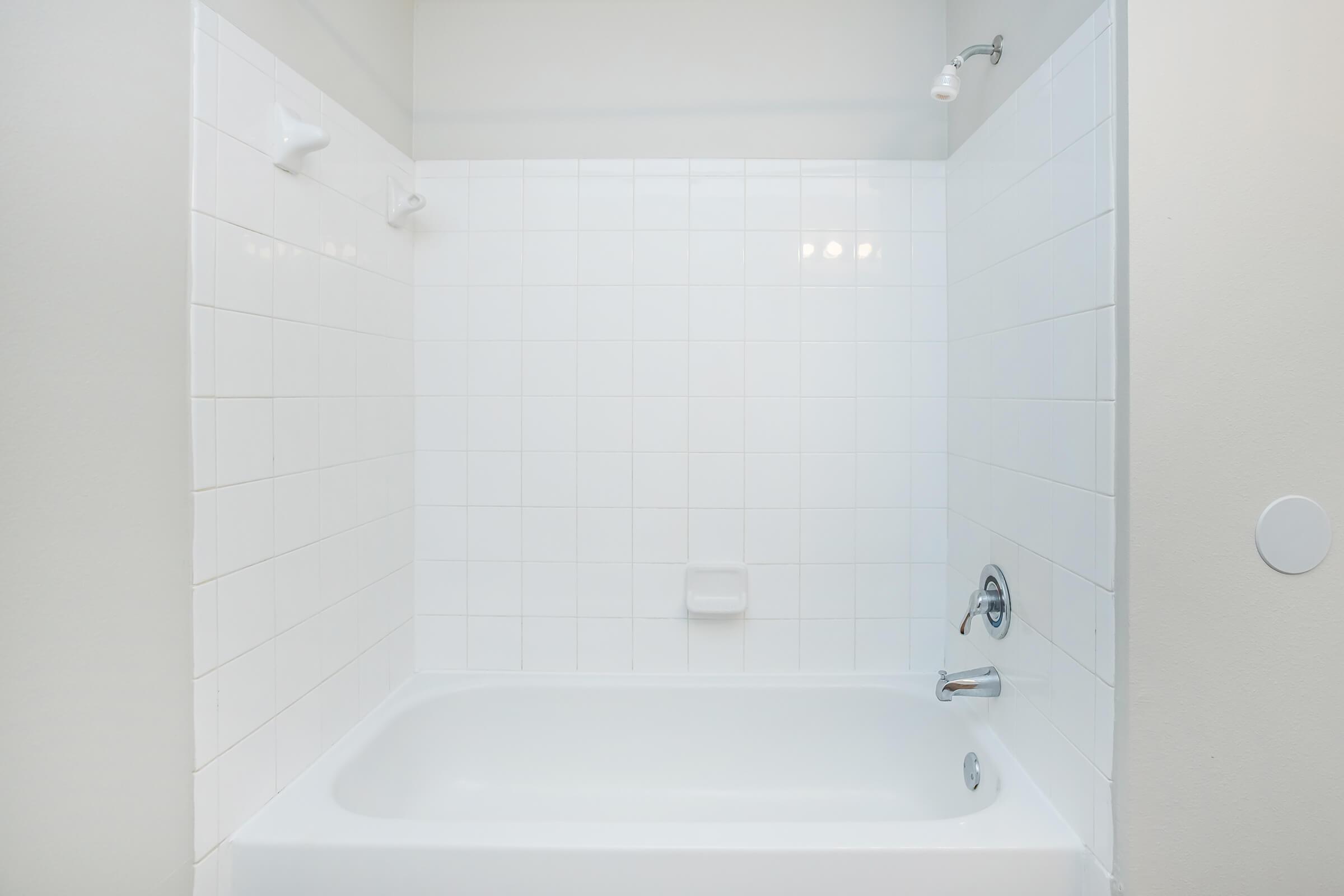 A clean, modern bathroom featuring a white tiled bathtub and walls. The tub has a silver faucet and a showerhead above. Bright lighting enhances the uncluttered space, making it appear fresh and inviting.