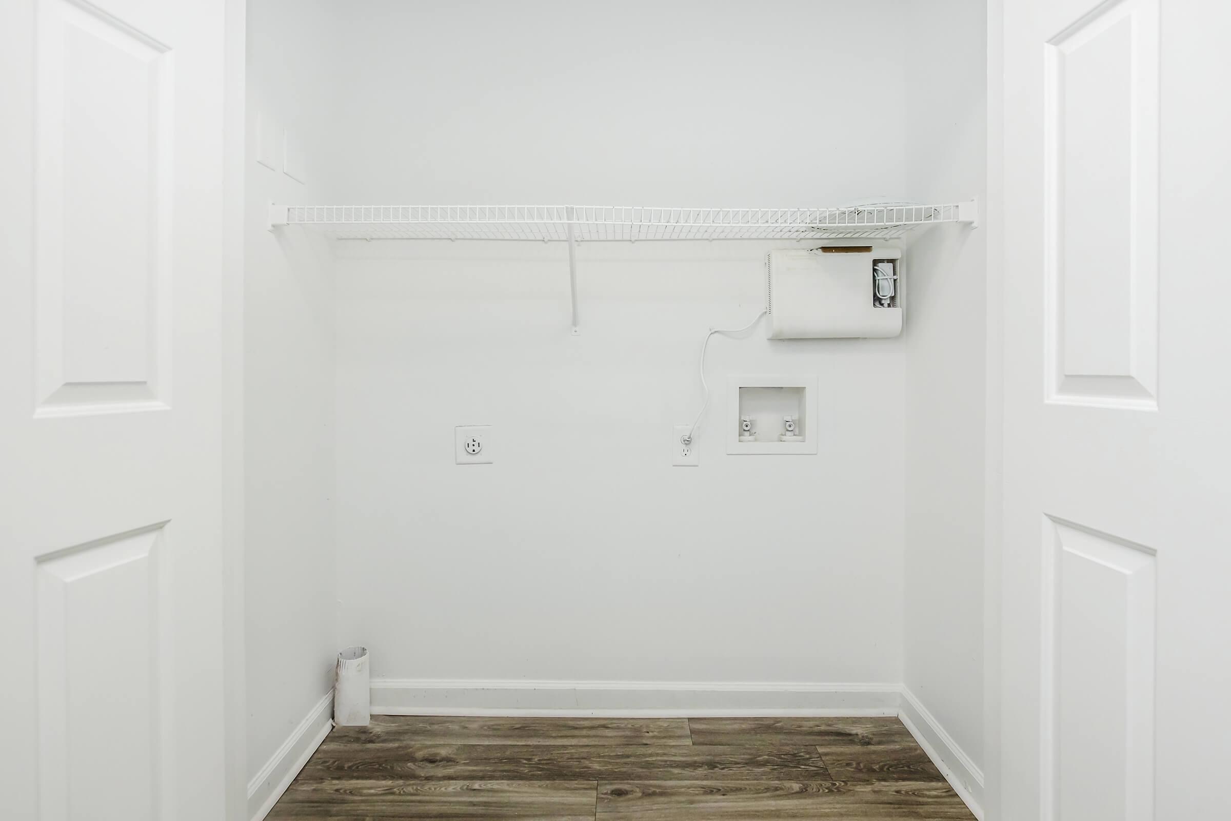An empty closet with white walls and a wooden floor. There is a wire shelf along the top, and wall outlets visible. No items are in the closet, creating a clean and spacious appearance.