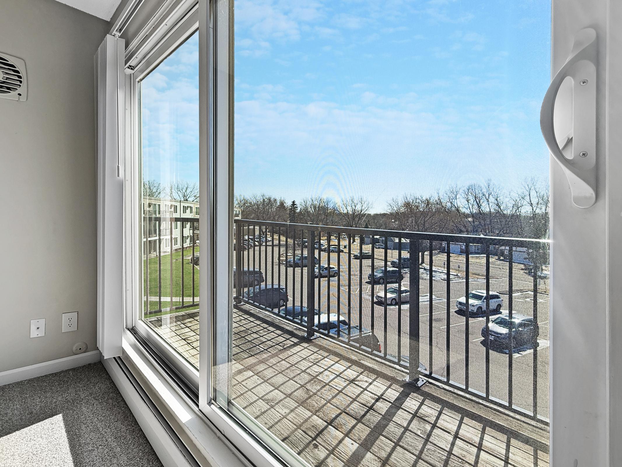 View from a balcony showcasing a parking lot filled with cars and green spaces beyond. The image captures a bright, sunny day with a clear blue sky and scattered clouds, framed by a large glass sliding door. The interior of the room features neutral-colored walls and carpet.