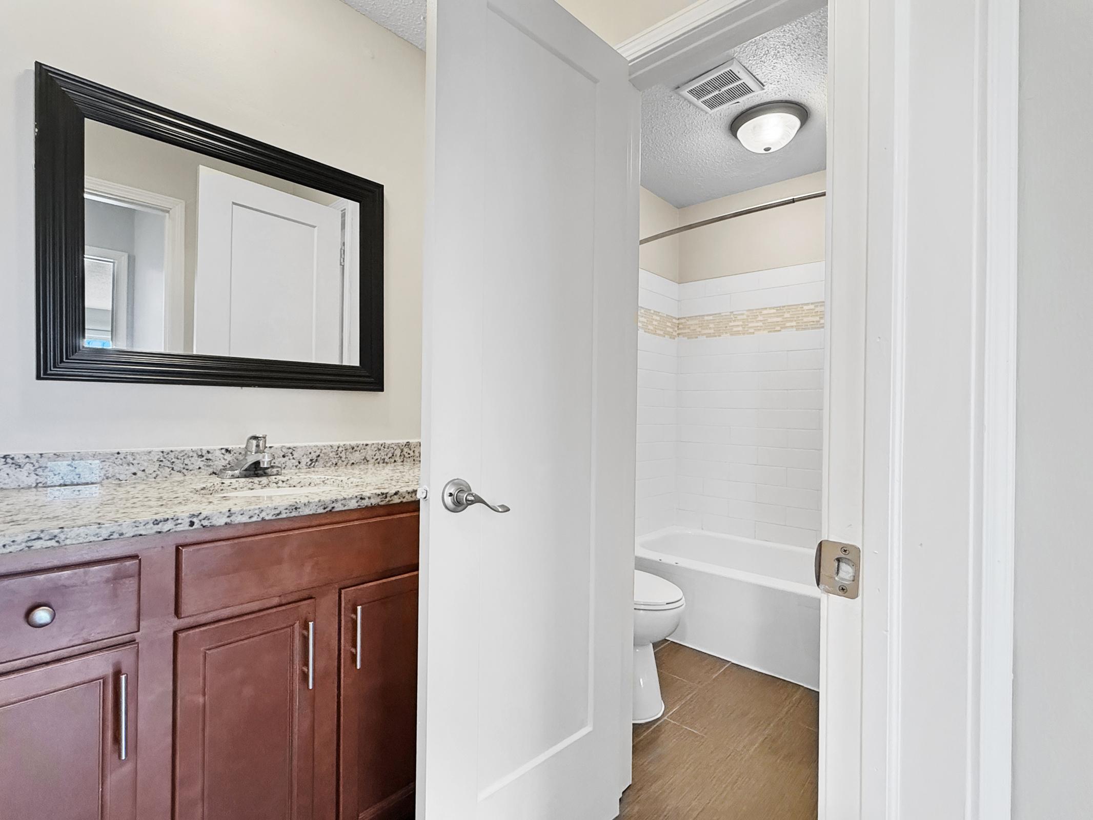 A view of a bathroom featuring a bathtub, toilet, and a granite countertop sink. The room has beige walls and a large mirror above the sink. A partially open door leads to another area, with a light fixture in the ceiling. The space is clean and well-lit.