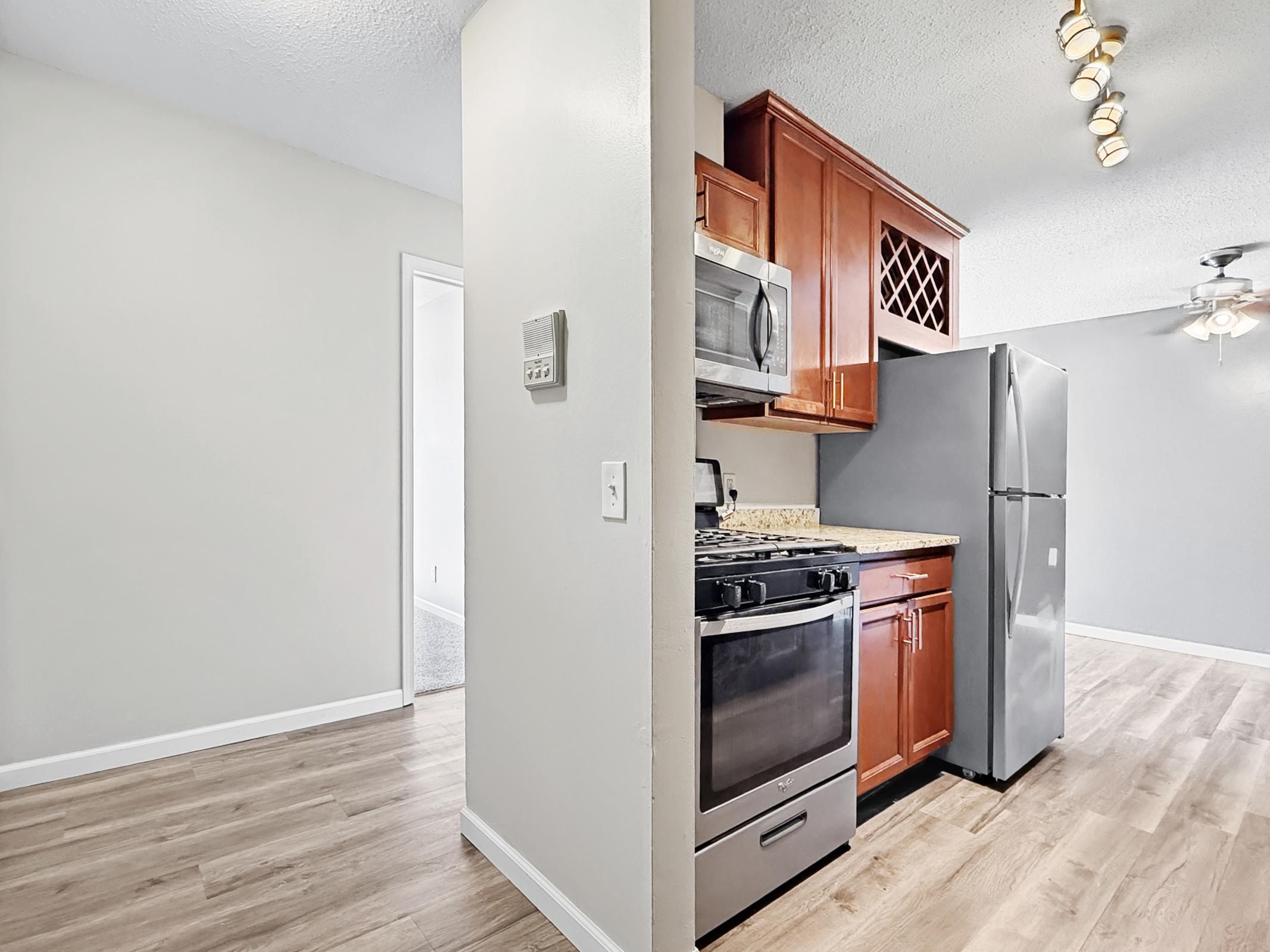 A modern kitchen featuring stainless steel appliances, including a gas stove and refrigerator. The cabinets are a rich wood tone, complemented by a granite countertop. Hardwood flooring extends into the adjacent living area, which has a ceiling fan and a neutral color scheme. The space is well-lit and inviting.