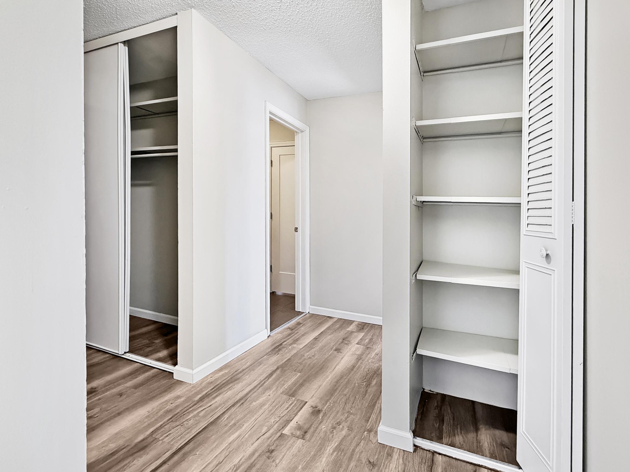 A well-lit room with light-colored walls and laminate flooring. On the left, there is a closet with sliding mirrored doors, and on the right, a shelving unit. In the background, a doorway leads to another room, creating an open and spacious feel.