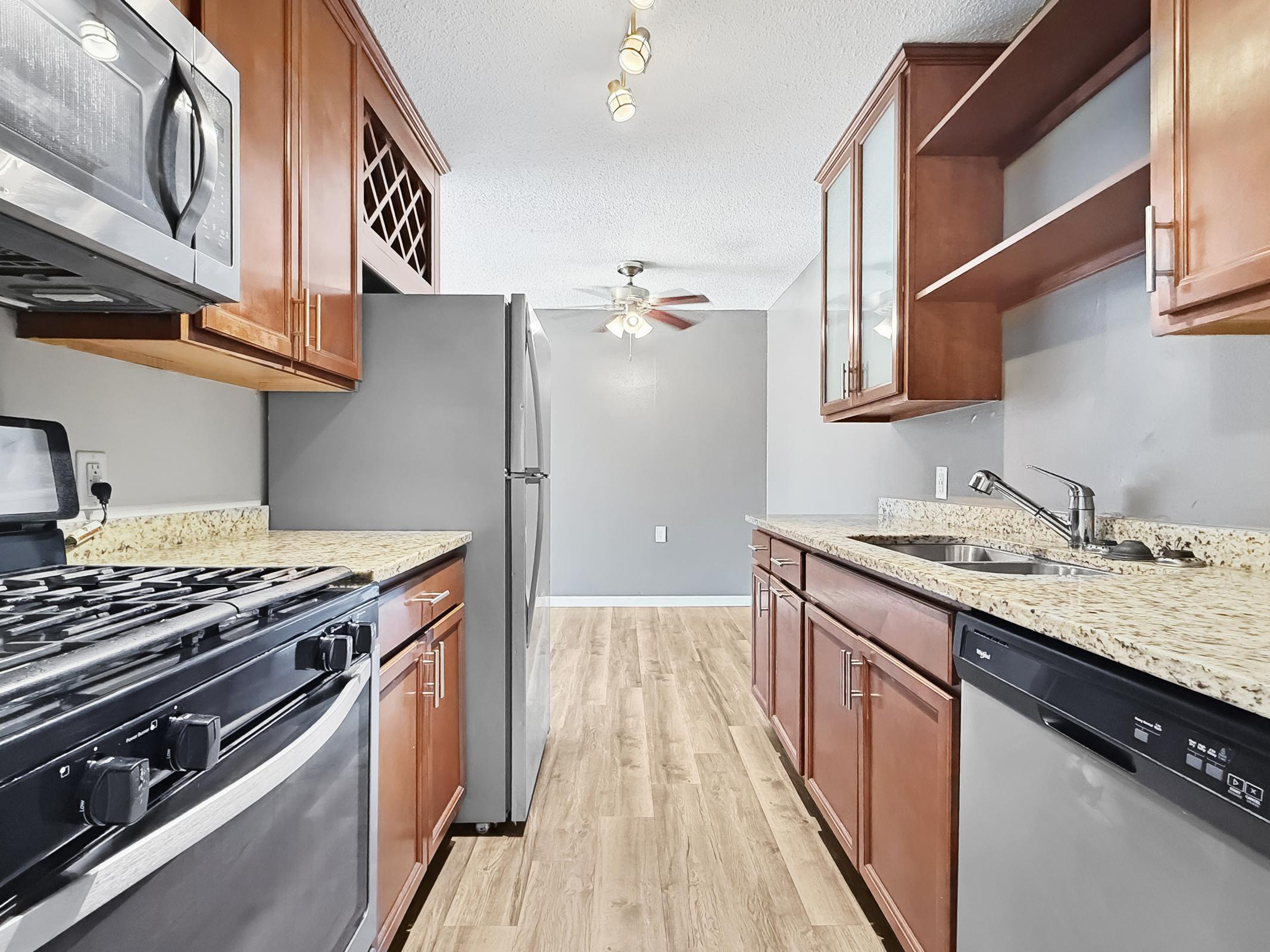 A modern kitchen featuring wooden cabinets, stainless steel appliances, and granite countertops. The layout includes a gas stove, refrigerator, and sink, with an open view to a light-filled living area. The walls are painted in a neutral gray, creating a contemporary atmosphere.