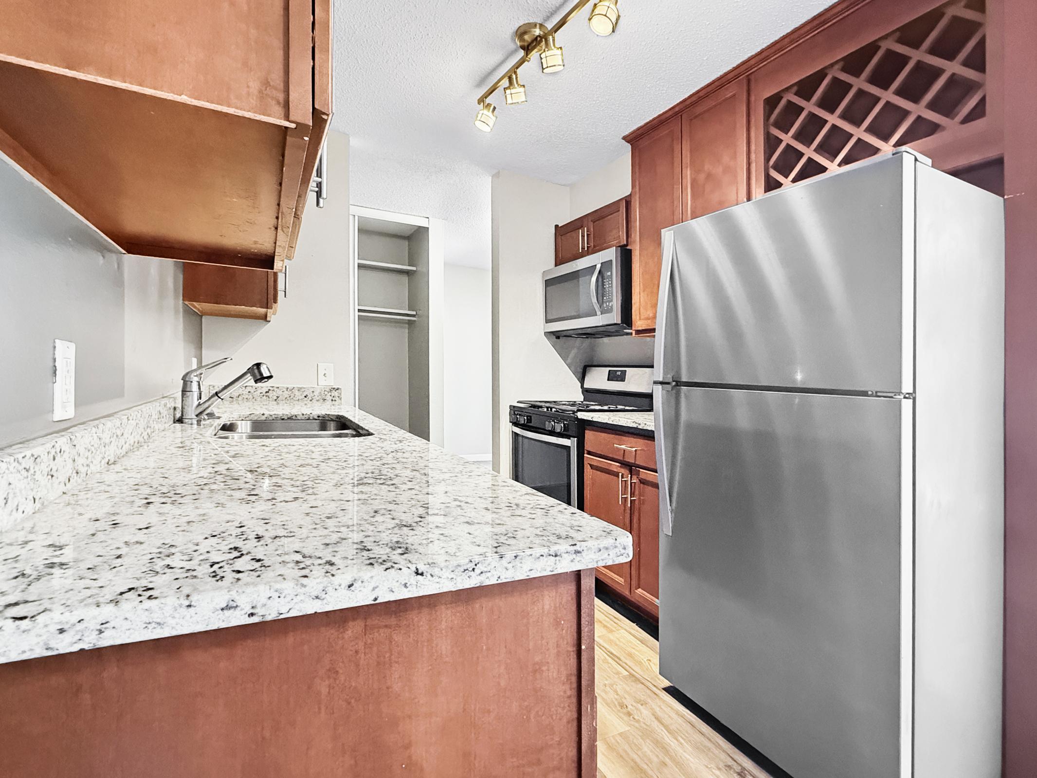 Modern kitchen featuring a granite countertop, stainless steel refrigerator, and stove. The cabinetry is wooden with a warm finish, and there is a wine rack above the sink. A spacious pantry area is visible in the background, with natural light illuminating the space.