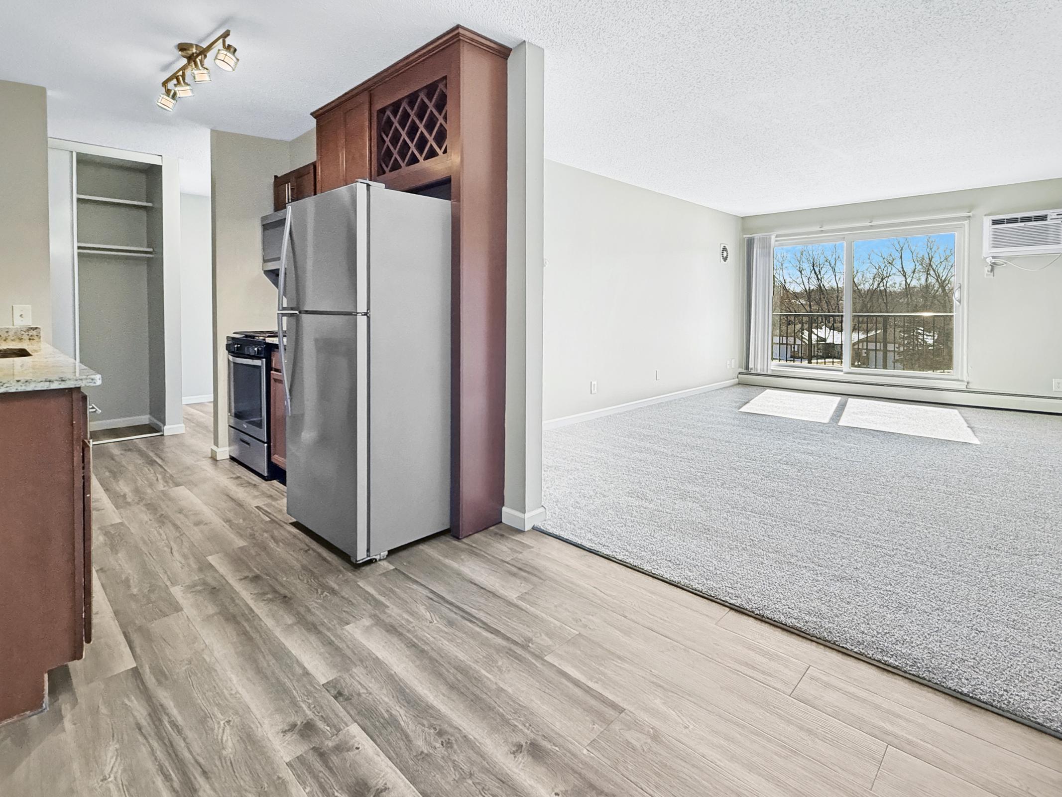 A bright, modern living space featuring a kitchen with stainless steel appliances and wood cabinets. The open layout leads to a spacious living area with large windows, providing plenty of natural light. Neutral walls and flooring create a clean and inviting atmosphere.