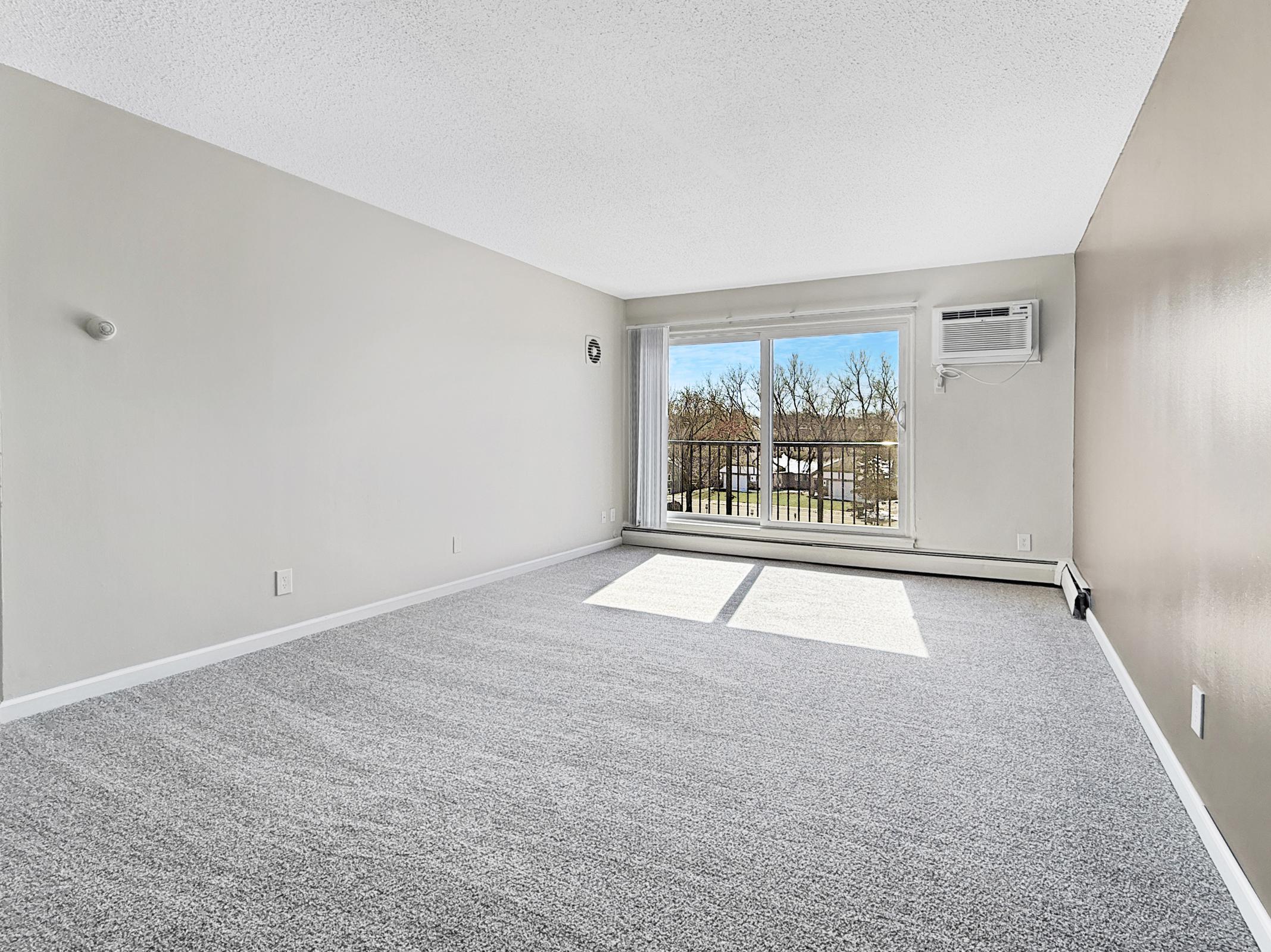 A spacious, vacant room with light gray carpet and a large window allowing natural light. The walls are painted in neutral colors, with a small wall clock. An air conditioning unit is mounted above the window, and there is a balcony visible outside the window.