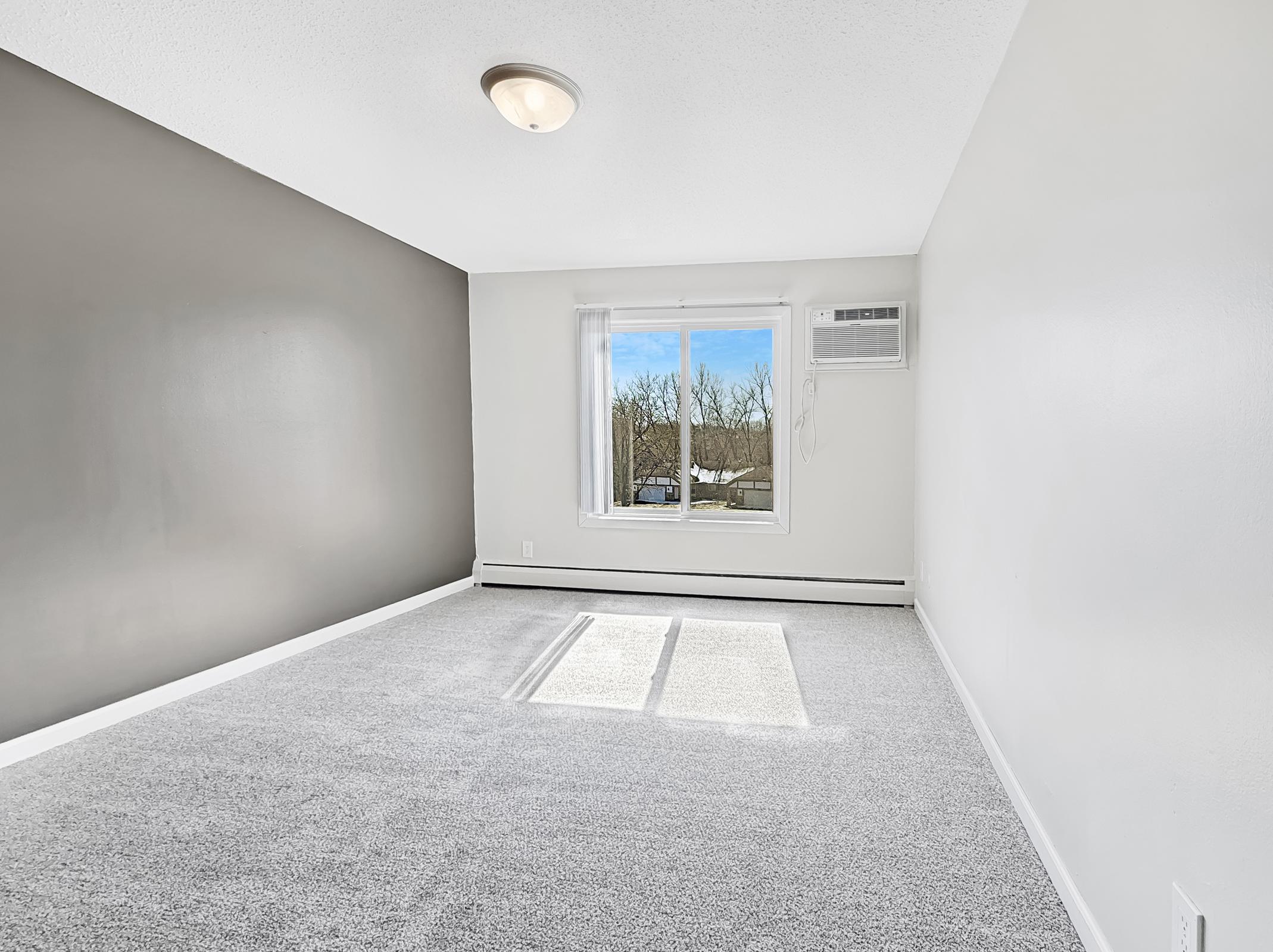 Empty room with light gray carpet and a two-tone wall featuring a dark gray section. A window lets in natural light, showing trees outside. An air conditioning unit is mounted above the window. The ceiling is plain white with a ceiling light fixture. Overall, the space appears bright and inviting.