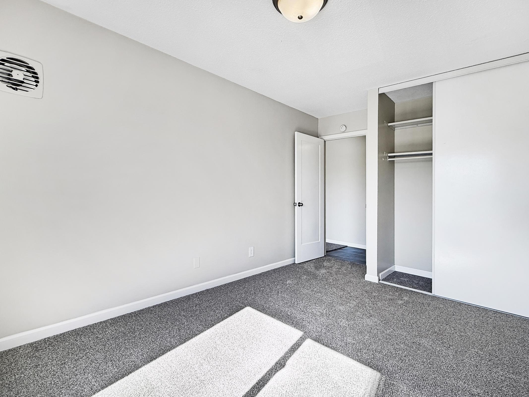 An empty, well-lit room with gray carpet and light gray walls. There is a closet with sliding doors on one side, and a white door leading to another space. The ceiling features a simple light fixture, and there is an air vent on the wall. Sunlight is casting shadows on the floor.