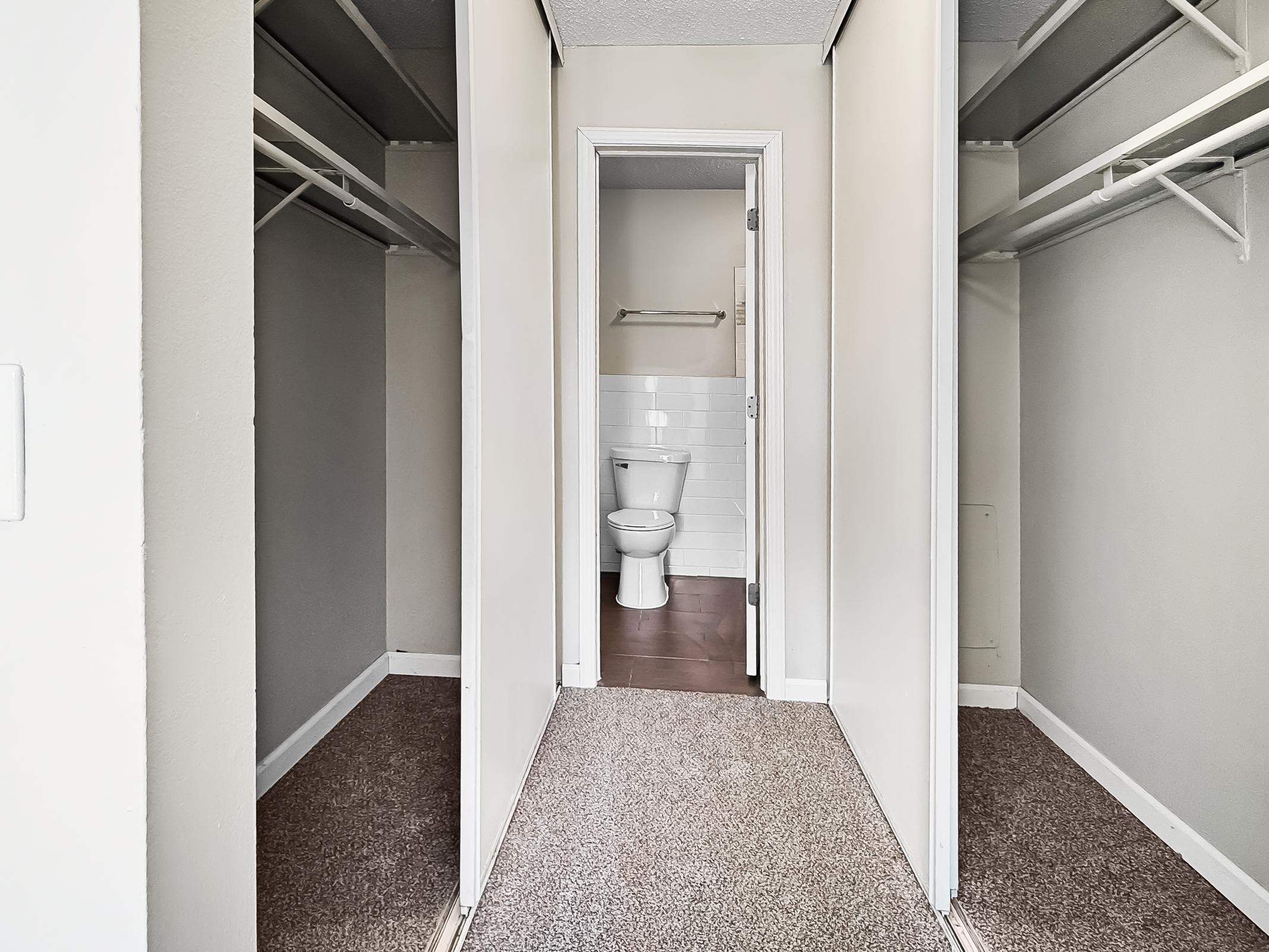 Interior view of a closet with sliding door panels, featuring a carpeted floor and shelves on either side. At the end of the closet, a bathroom is visible, showing a white toilet and partial wall tiling. The walls are painted in light colors, creating a spacious and organized look.