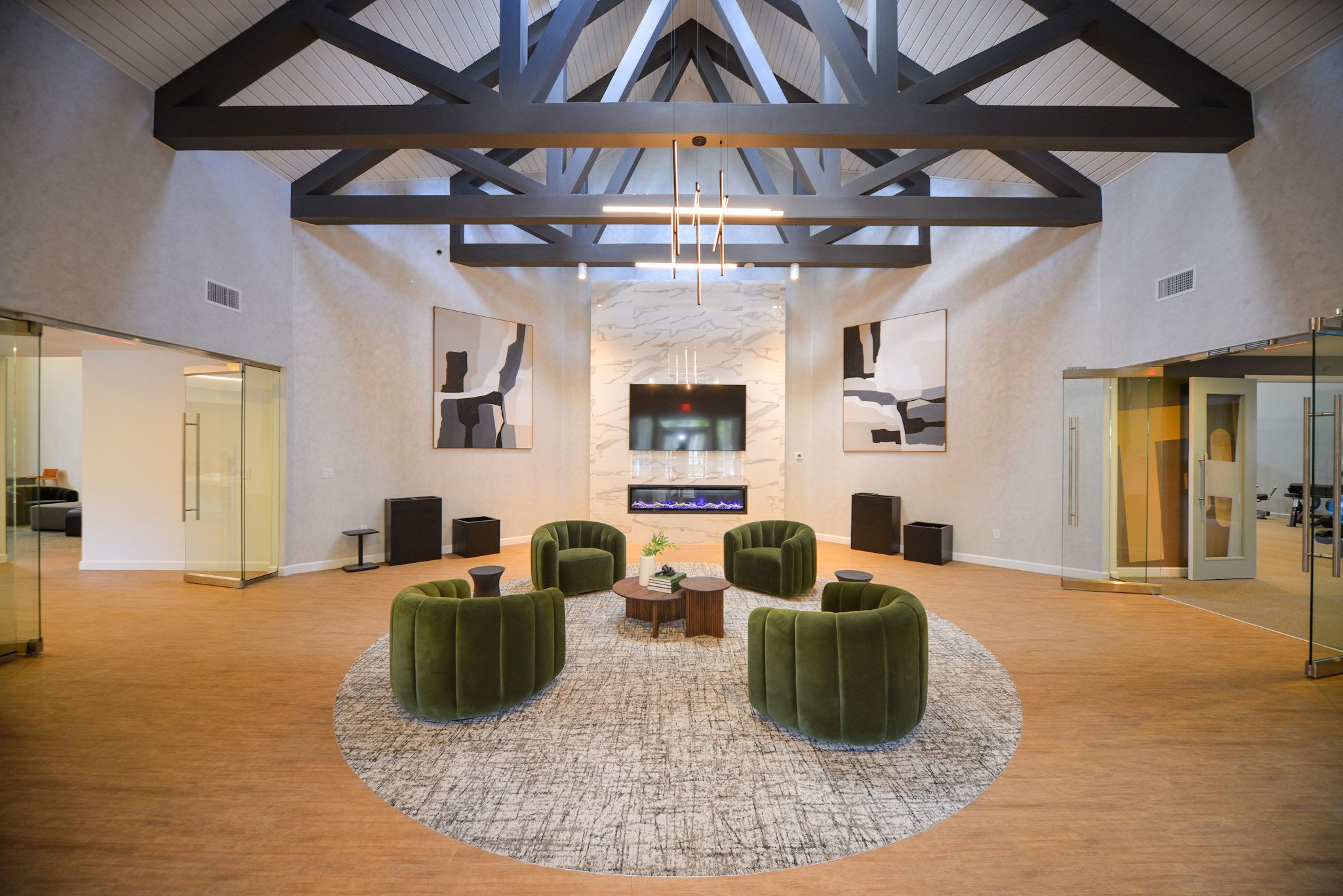 A modern lobby featuring a circular layout with green velvet armchairs arranged around a circular area rug. A large flat-screen TV is mounted on a textured wall. Abstract artwork hangs on the walls, and there are glass doors leading to other spaces. The ceiling has exposed beams, creating a spacious and contemporary atmosphere.